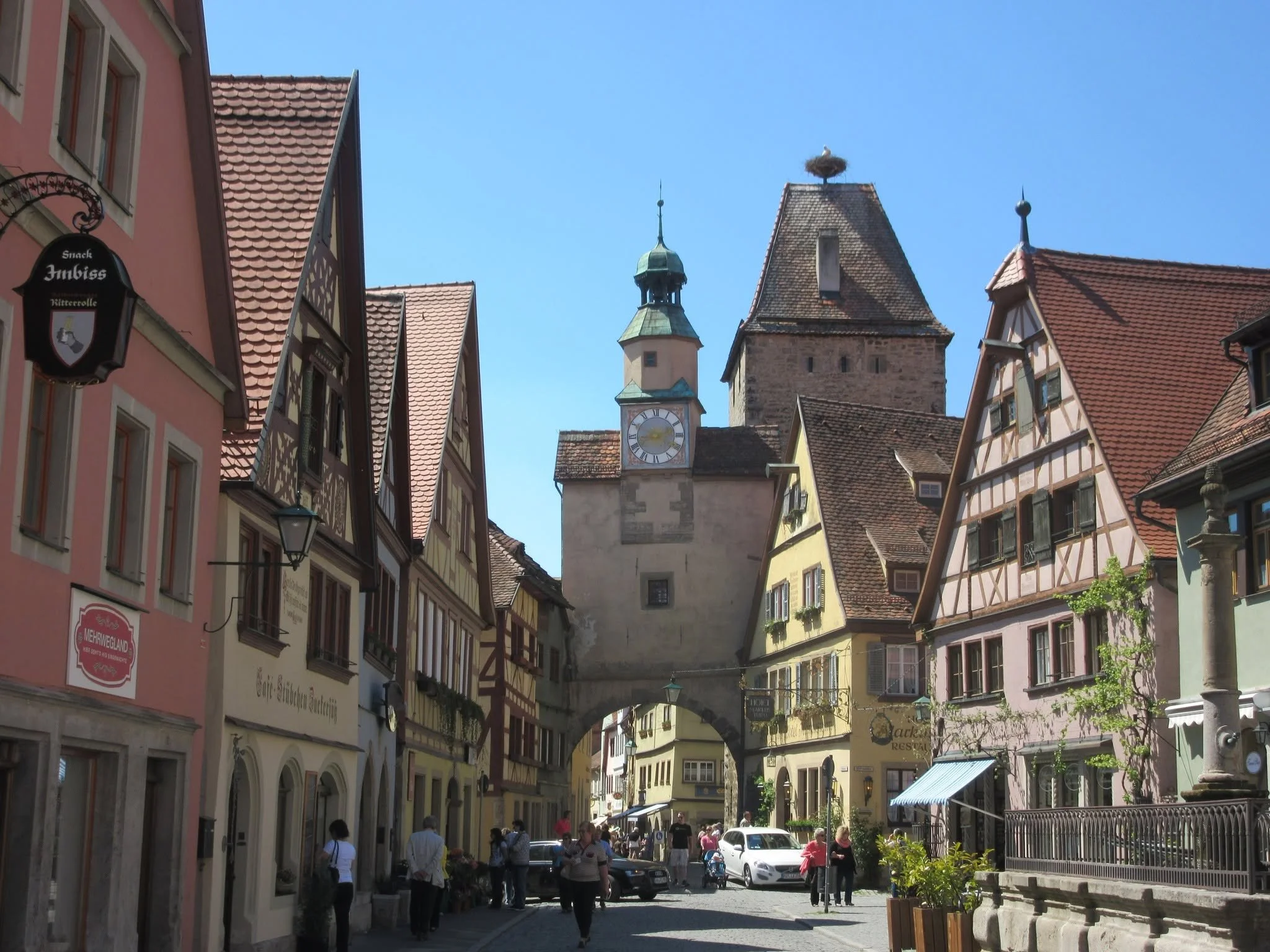A street scene in a European town with historic buildings, a clock tower, stork nest, and people walking.