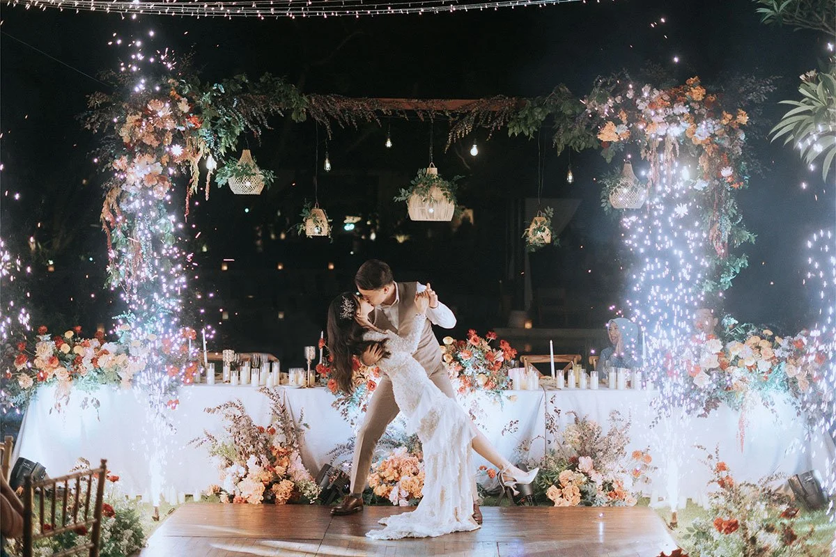 A newlywed couple is dancing and kissing at their wedding reception, with fireworks and floral decorations surrounding them.
