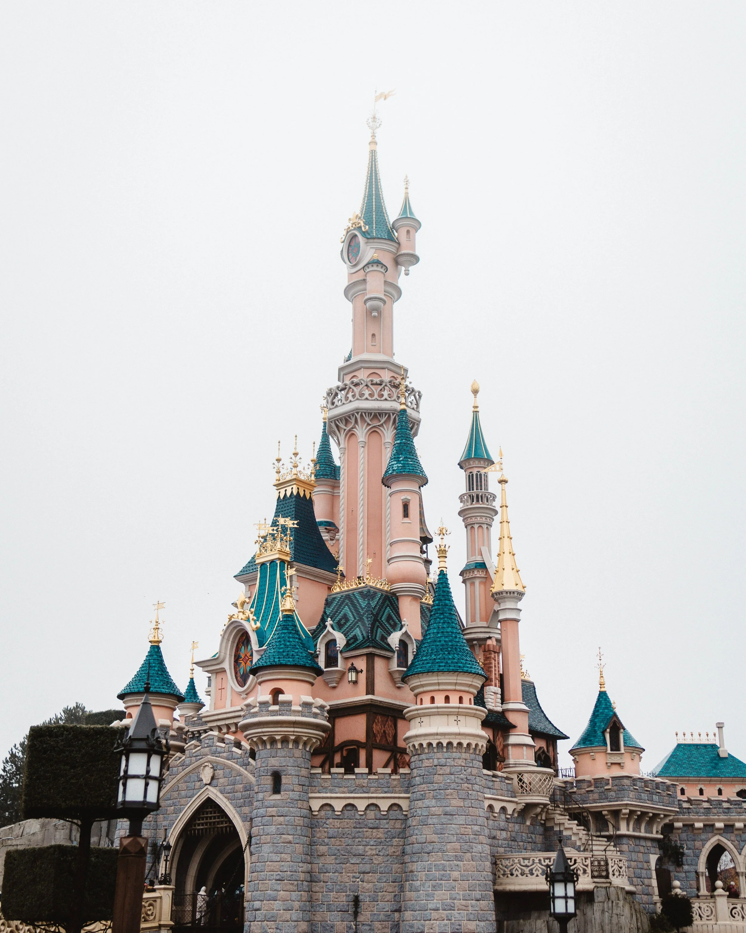 Castle with pink and blue turrets, stone walls, decorative spires, and black lanterns in a theme park.