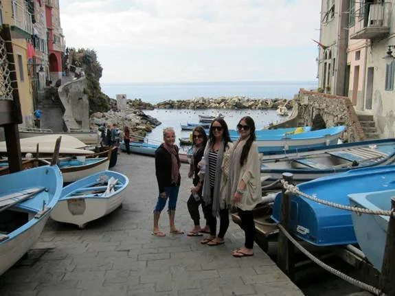 Four women pose on a cobblestone street near small blue and white boats with a rocky shoreline and the sea in the background.