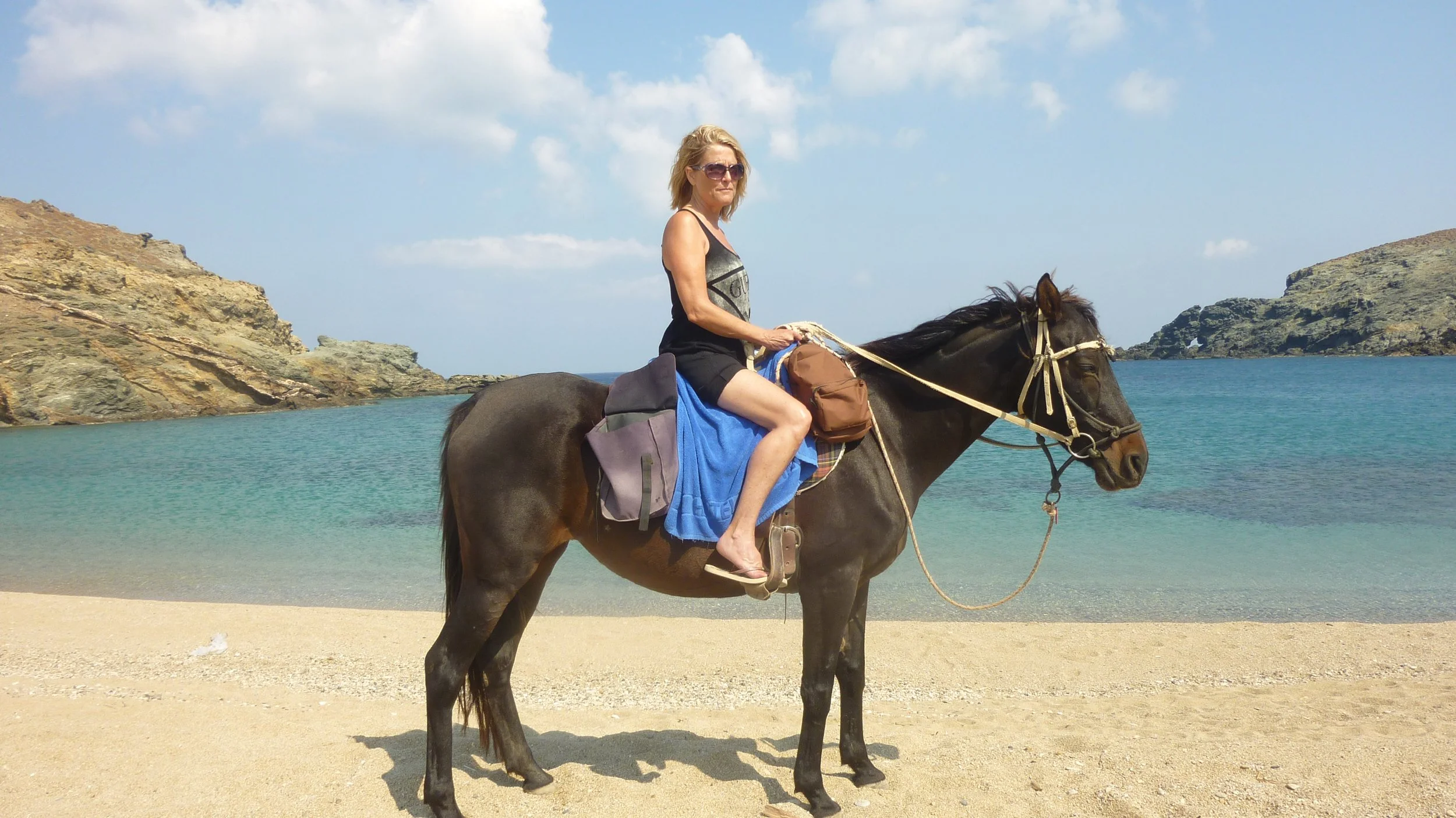 A woman riding a horse on a beach near a body of water with rocky cliffs in the background.