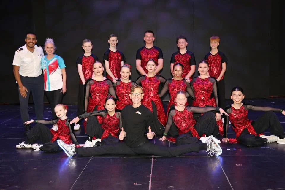 A group of young girls in red and black costumes poses on stage with two adults, one man in a white shirt and another woman in black, all smiling.