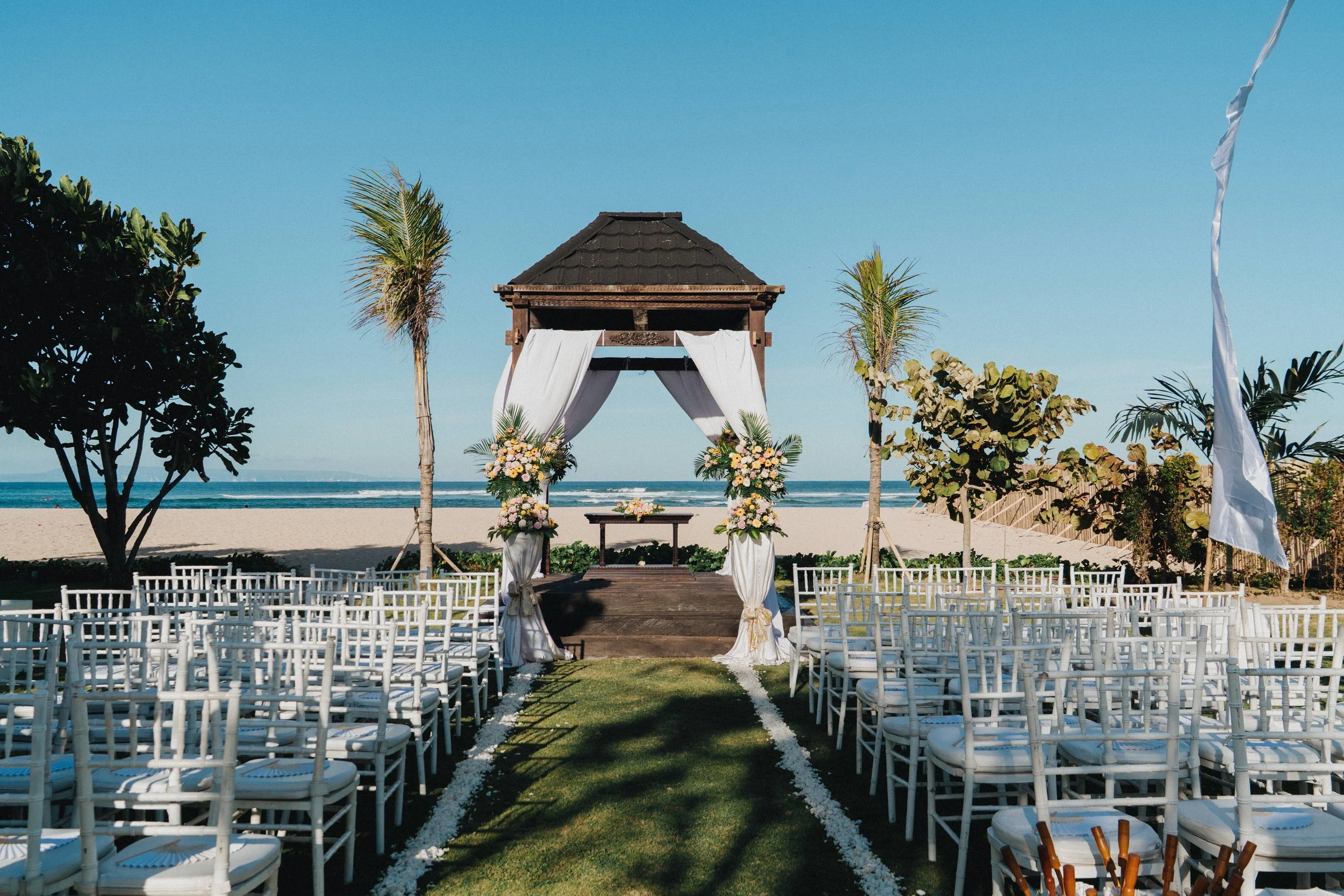 Beach wedding setup with white chairs, a wooden arch decorated with white curtains and flowers, on the sand by the ocean, under a clear blue sky.