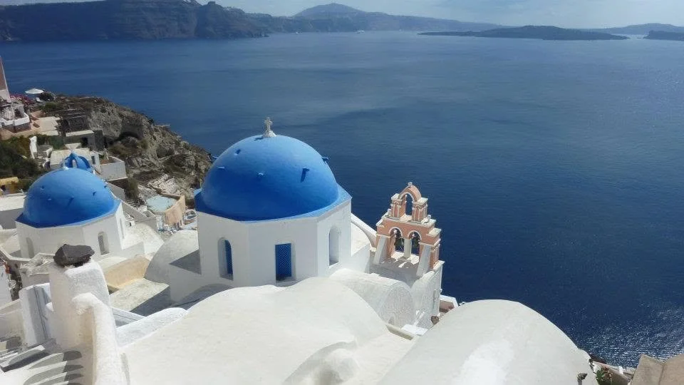 View of white buildings with blue domes and a church bell tower overlooking the deep blue sea in Santorini, Greece.