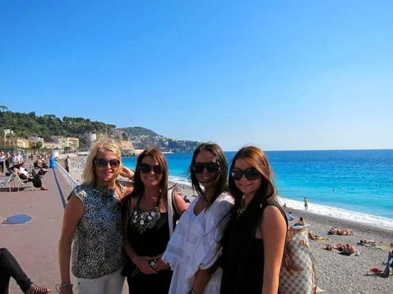 Four women standing together on a beach with the ocean and a shoreline in the background.