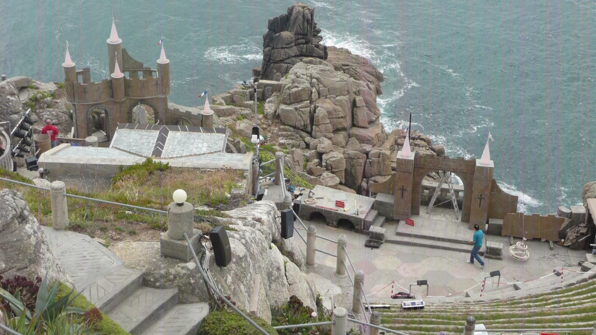 A stage set up near rocky cliffs and the sea with structures resembling a castle and a bridge, some people working on the setup, and equipment like speakers and cables visible.