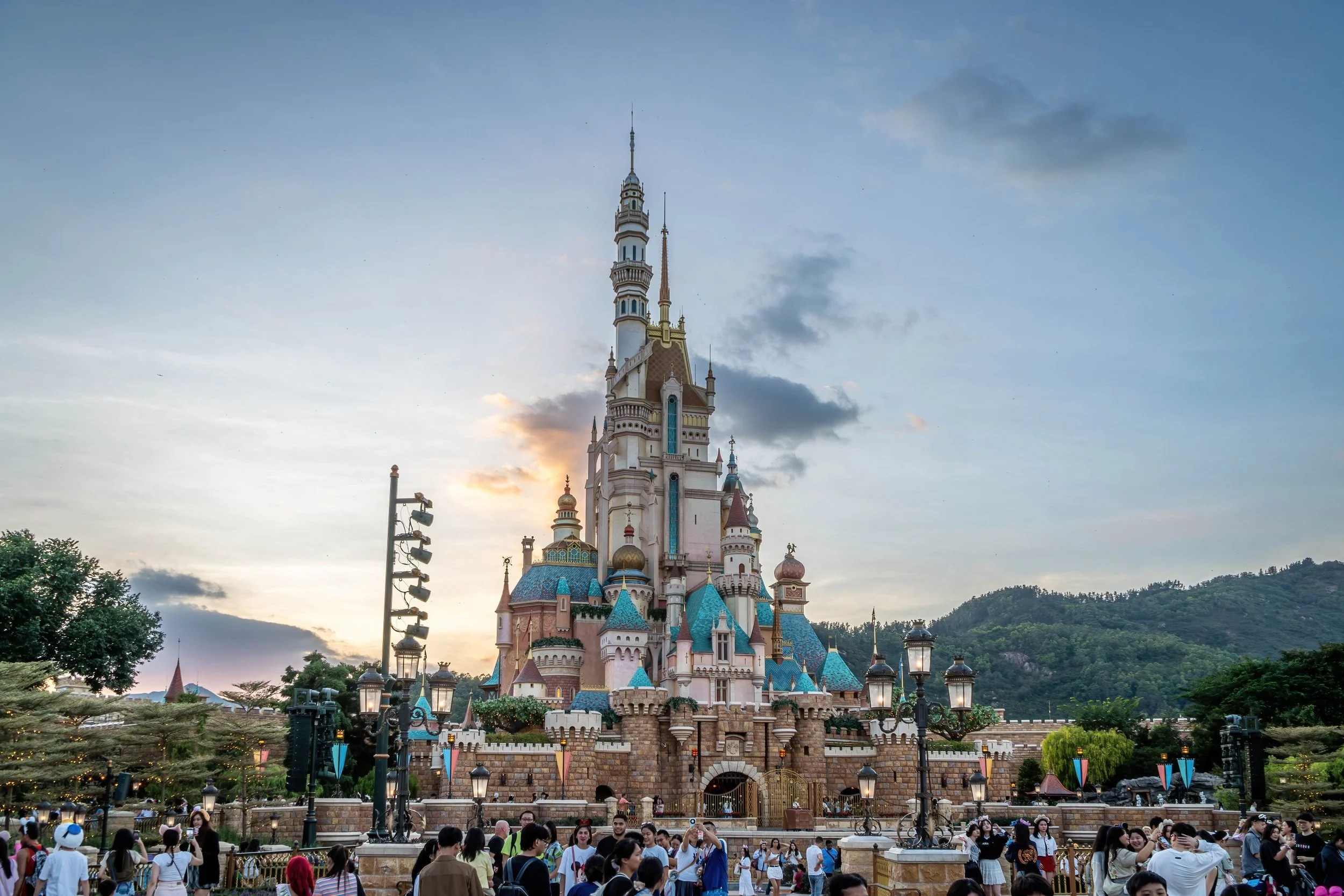 Castle at Disneyland with blue rooftops, surrounded by visitors and trees, with mountains in the background and a partly cloudy sky at sunset.