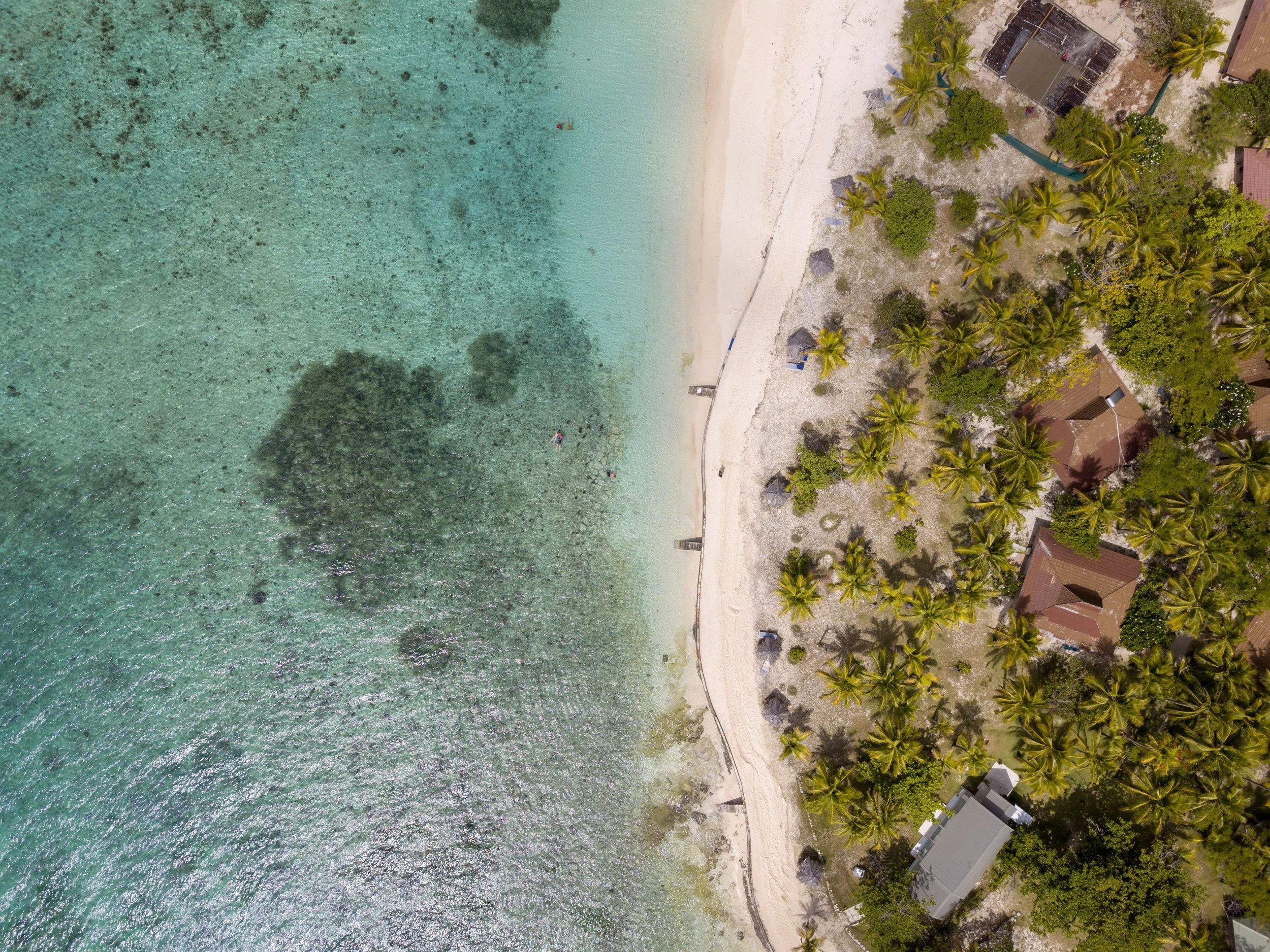 An aerial view of a beach with white sand, turquoise waters, and palm trees, along with some buildings and structures near the shoreline.