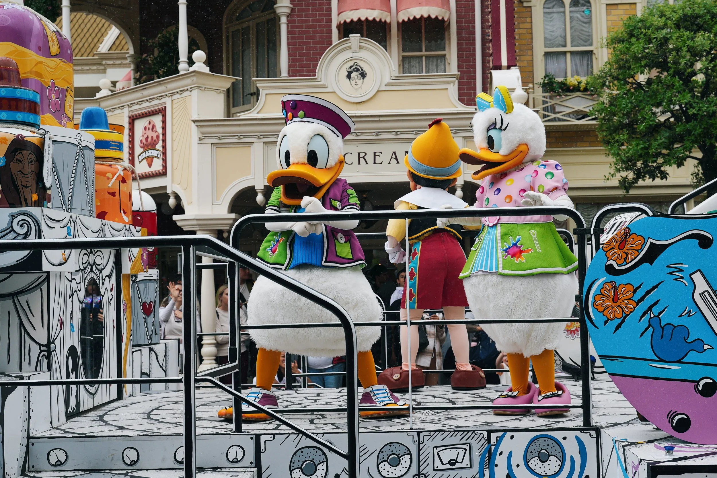 Costumed characters of Donald Duck and Daisy Duck on a parade float at a theme park, with people watching below. Donald is wearing a purple hat and jacket, while Daisy is dressed in a pink and green polka dot dress with a bow on her head.