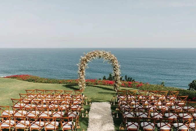 Wedding setup on a grassy hill overlooking the ocean with chairs arranged on either side of an aisle, and a floral arch at the end of the aisle.