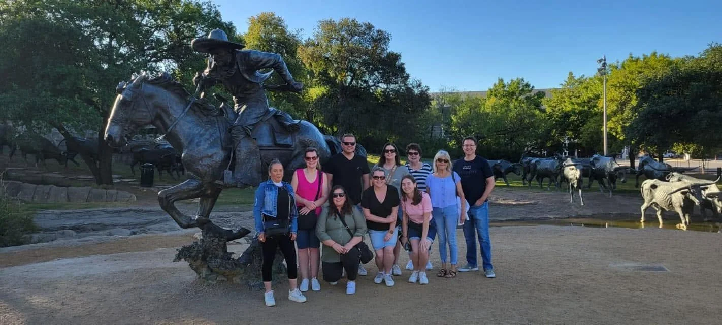 Group of nine people posing in front of a large cowboy statue on horseback at a park with trees and other animal statues in the background.
