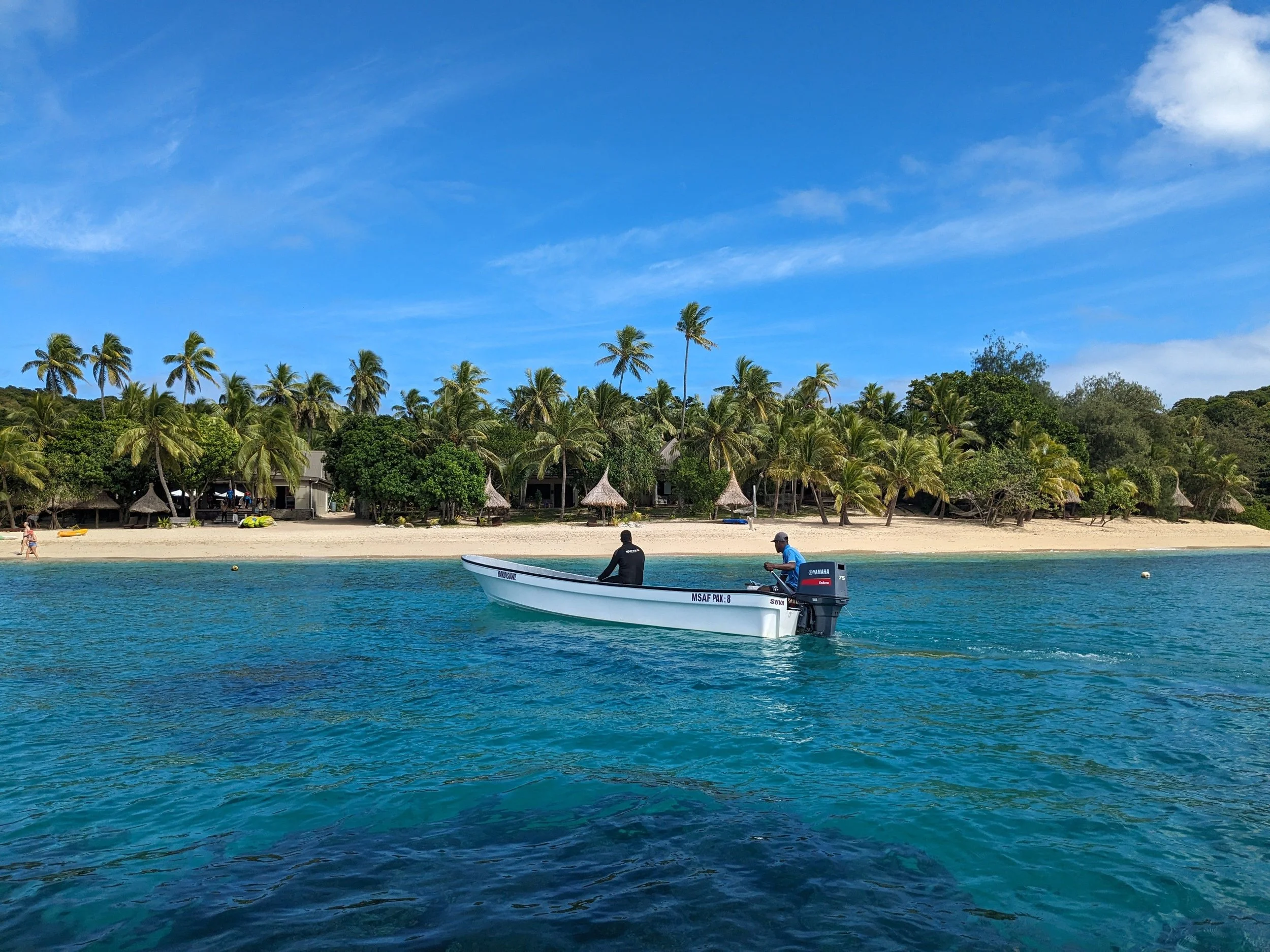 A boat with two people on it floating on clear blue water near a sandy beach lined with palm trees and thatched huts under a partly cloudy sky.