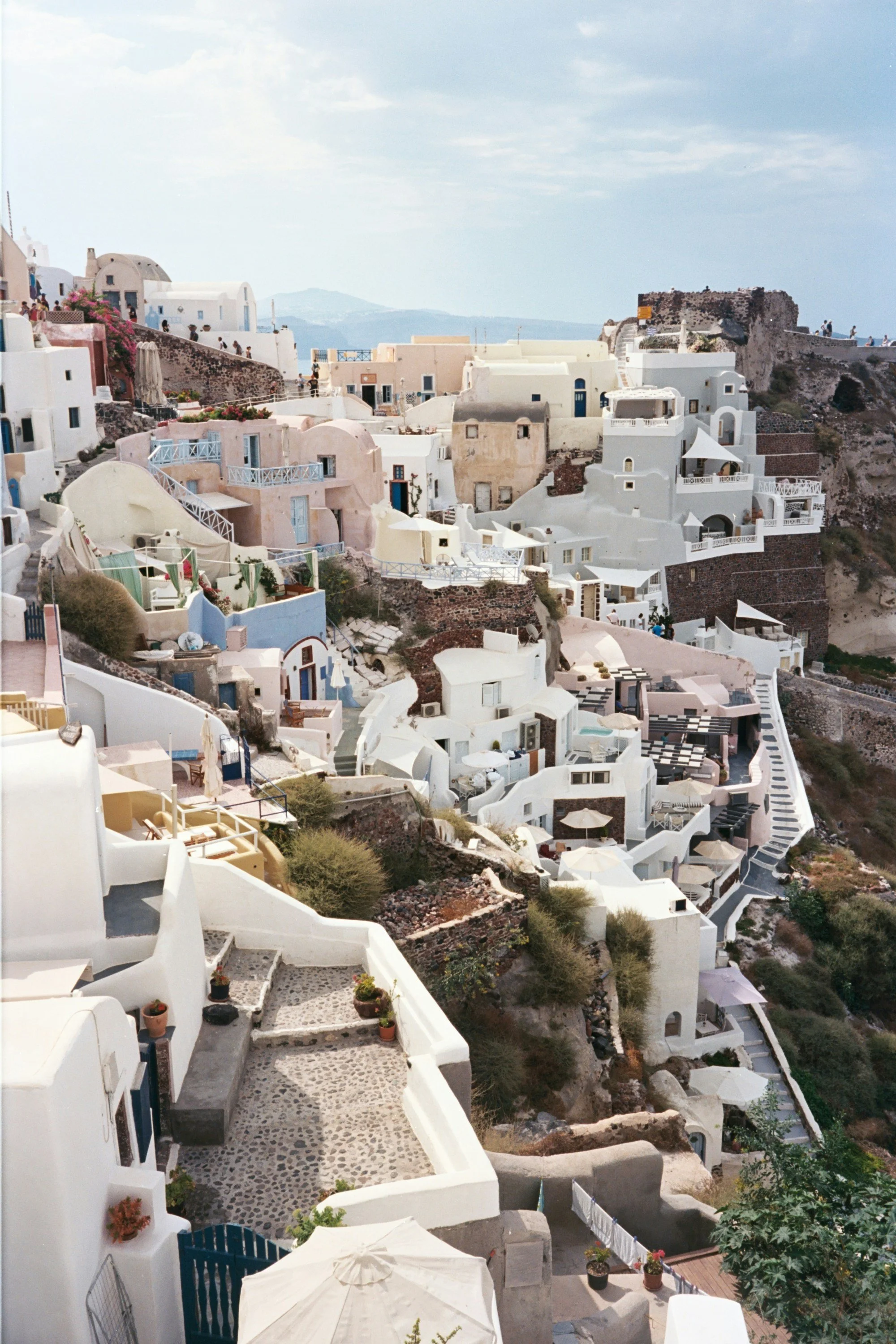 White and pastel-colored buildings with blue accents built into a hillside, typical of Santorini, Greece, under a partly cloudy sky.