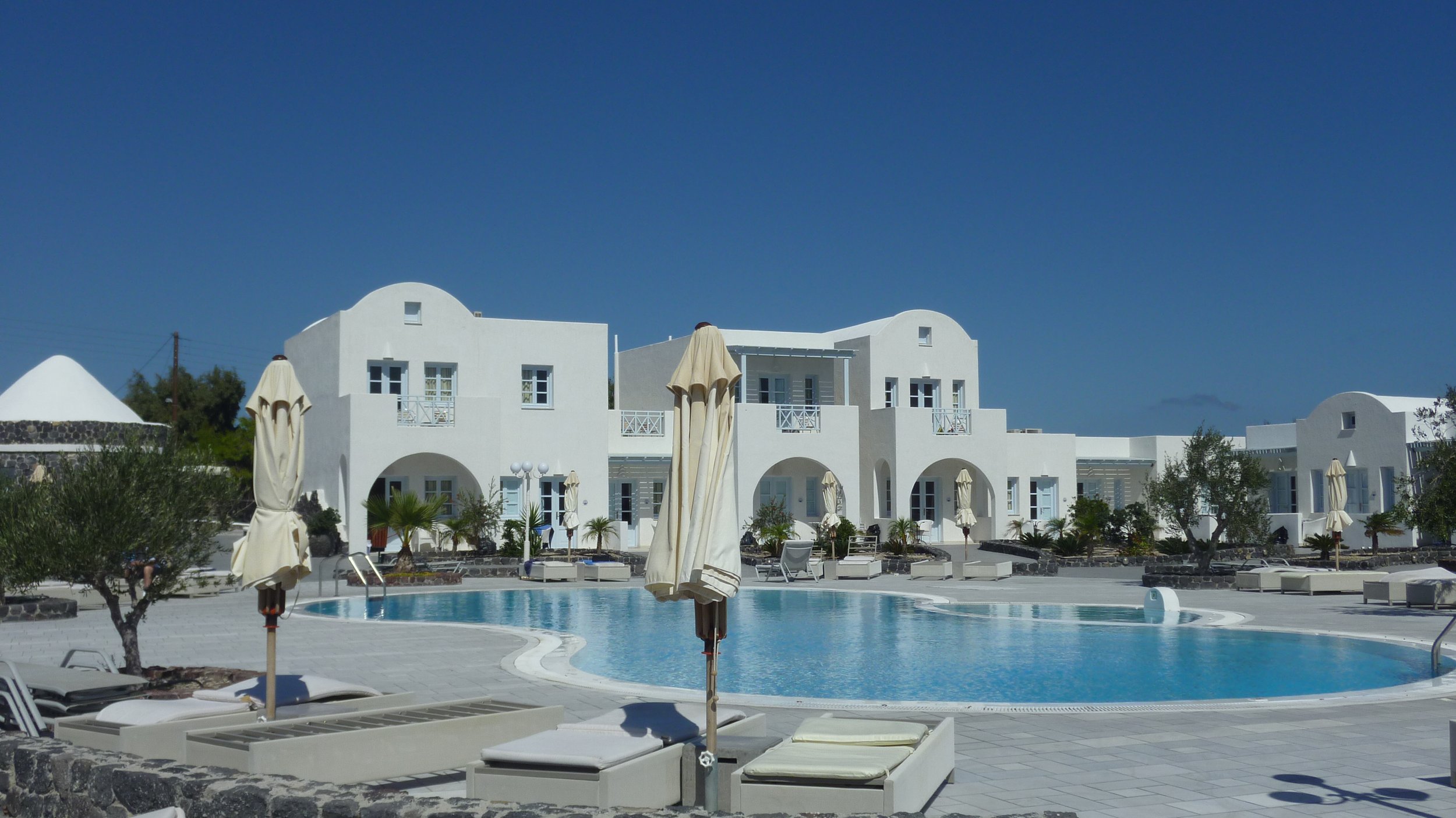 White Mediterranean-style hotel buildings with arches and balconies, swimming pool with lounge chairs and umbrellas, and lush greenery under a clear blue sky.