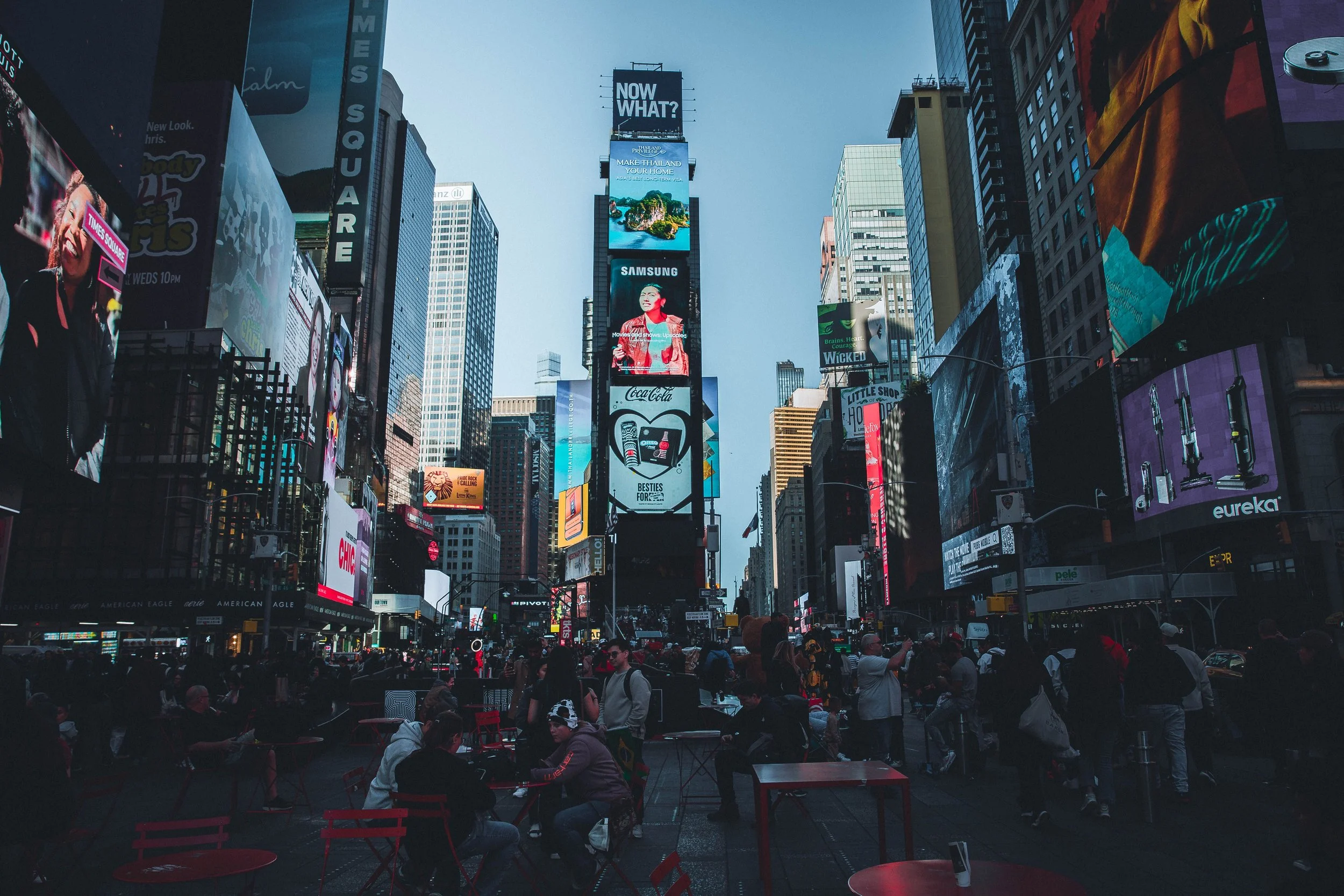 A busy city square filled with people sitting at red tables and chairs under tall buildings with large electronic billboards and advertisements.