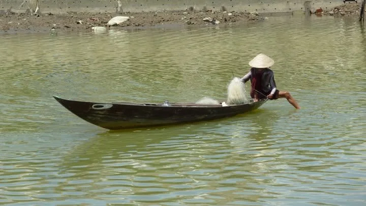 A person wearing a conical hat sitting in a small wooden boat on a calm body of water, with a fishing net.