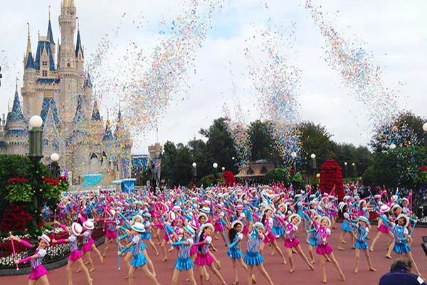 Large group of young girls dressed as cheerleaders in pink and blue costumes with pom-poms, performing at a Disney theme park in front of Cinderella's Castle, with confetti and streamers in the air.