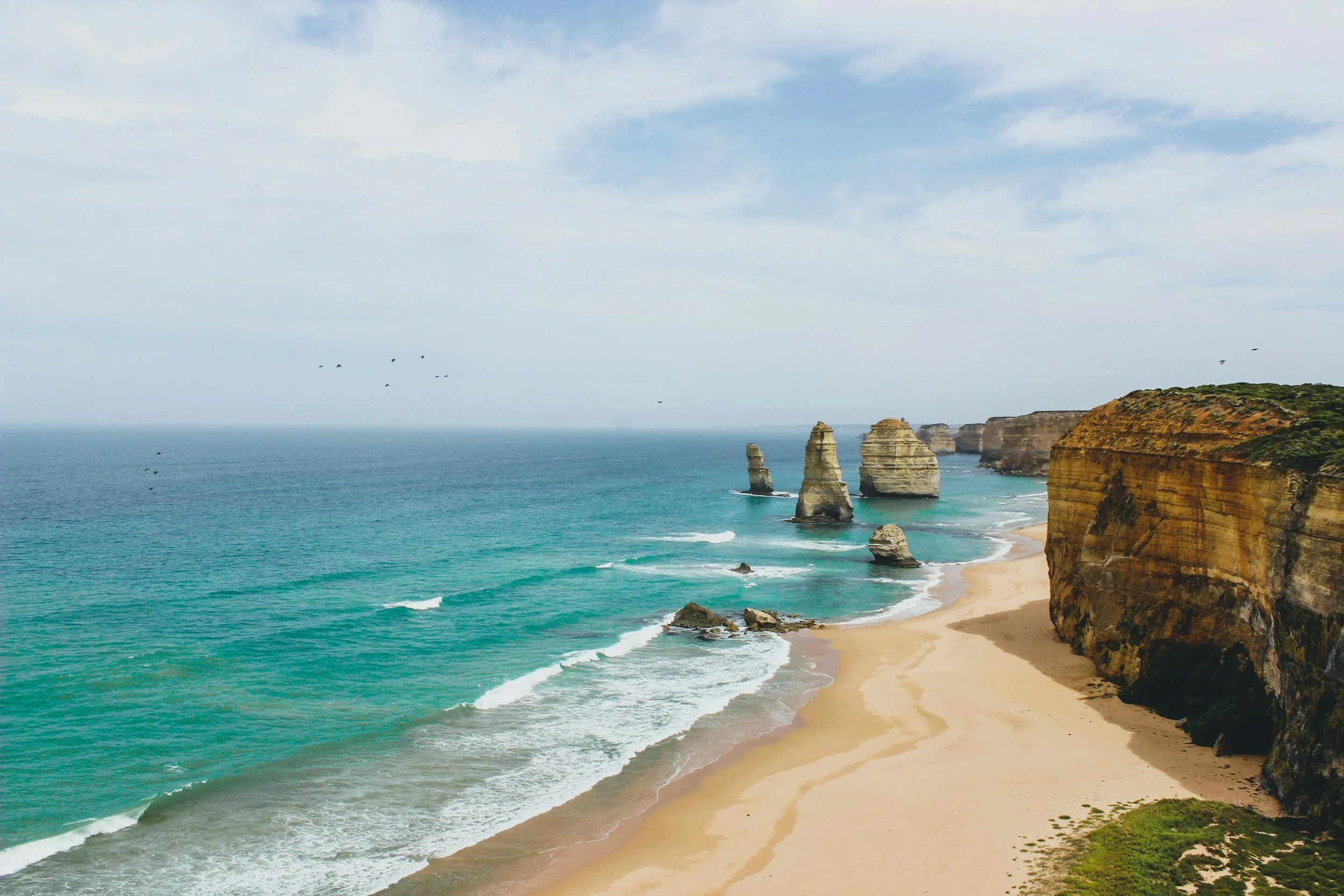 A scenic coastal landscape with turquoise ocean water, white cliffs, and rock formations. The sky is partly cloudy, and there are birds flying over the sea.