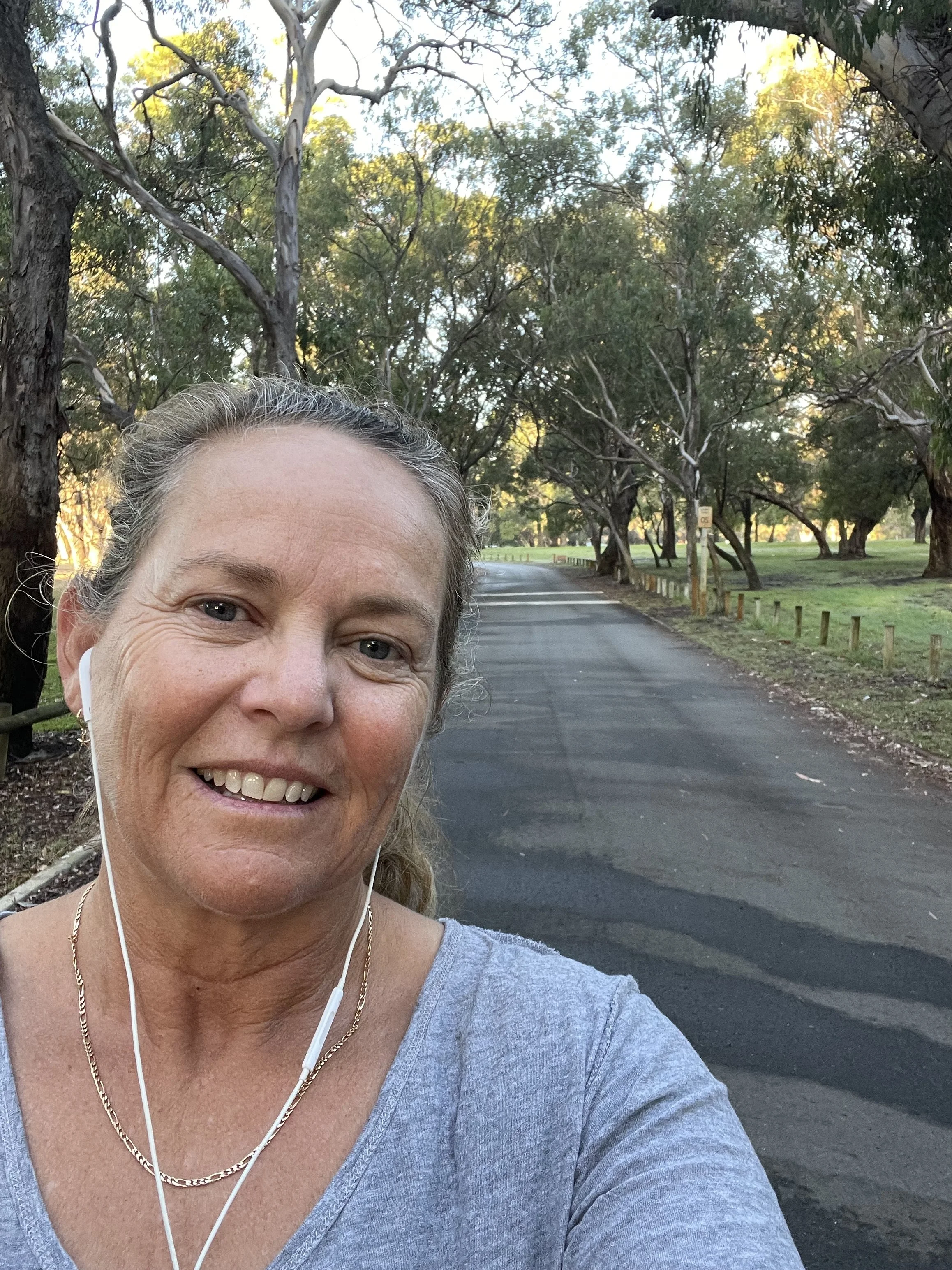 A woman smiling and wearing headphones taking a selfie in a park with tall trees and a paved path.