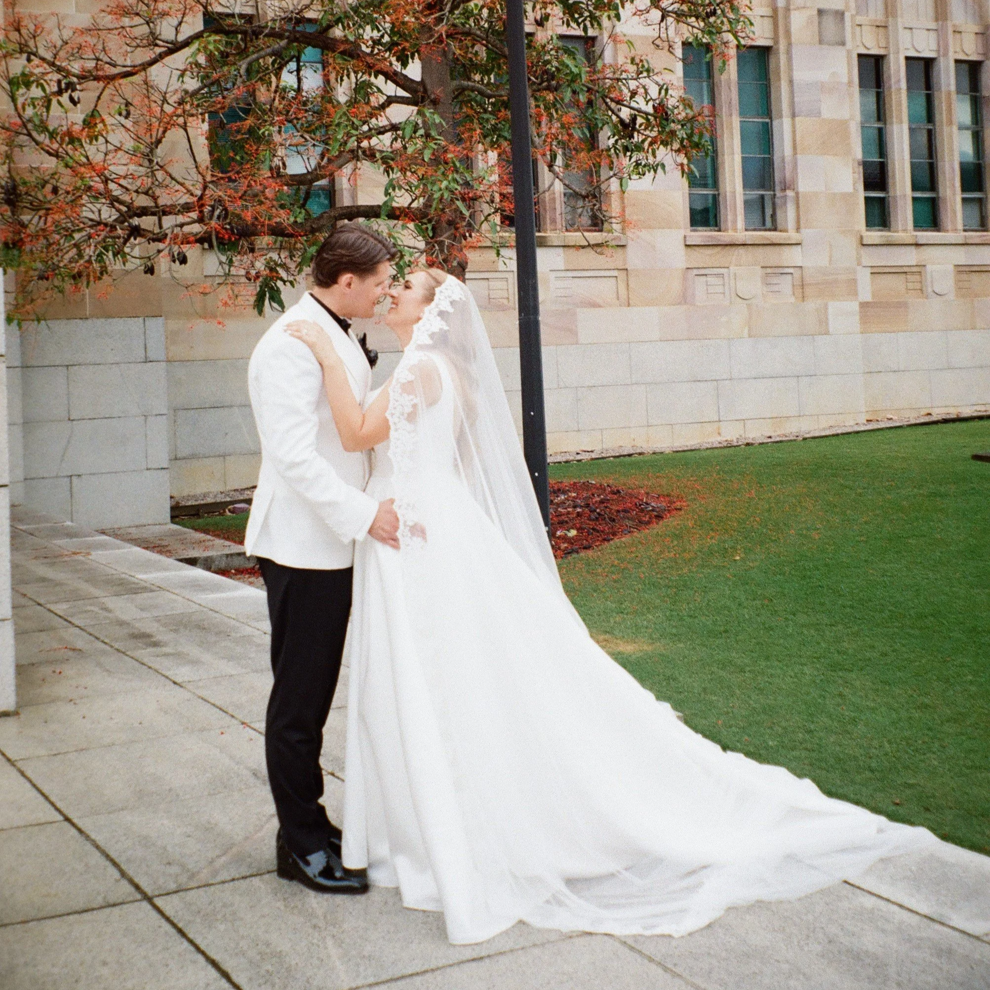 A bride and groom in wedding attire sharing a kiss outdoors near a building with beige stone walls and tall windows, with autumn trees nearby.