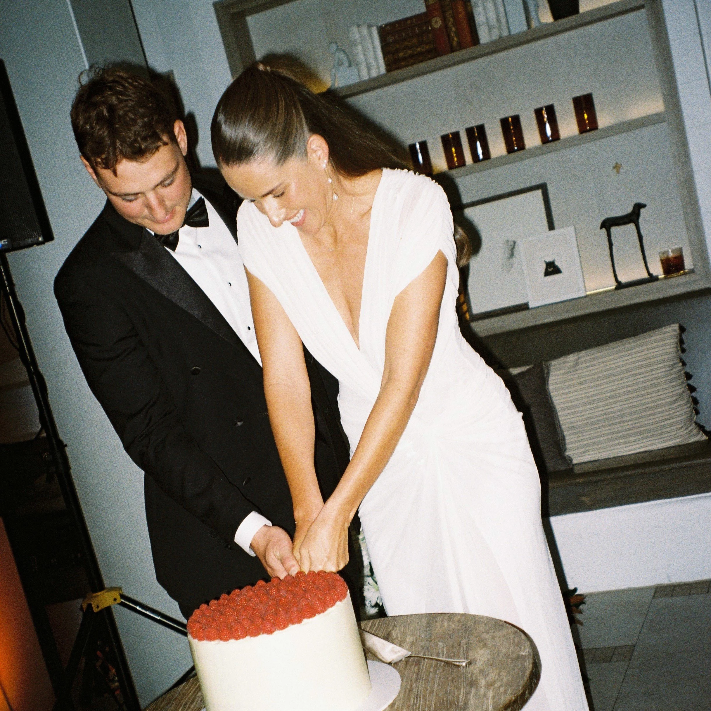 A couple dressed in formal attire, with the man in a black tuxedo and the woman in a white dress, leaning over a large white birthday cake decorated with strawberries on top, in a room with a bookshelf and decorative items in the background.