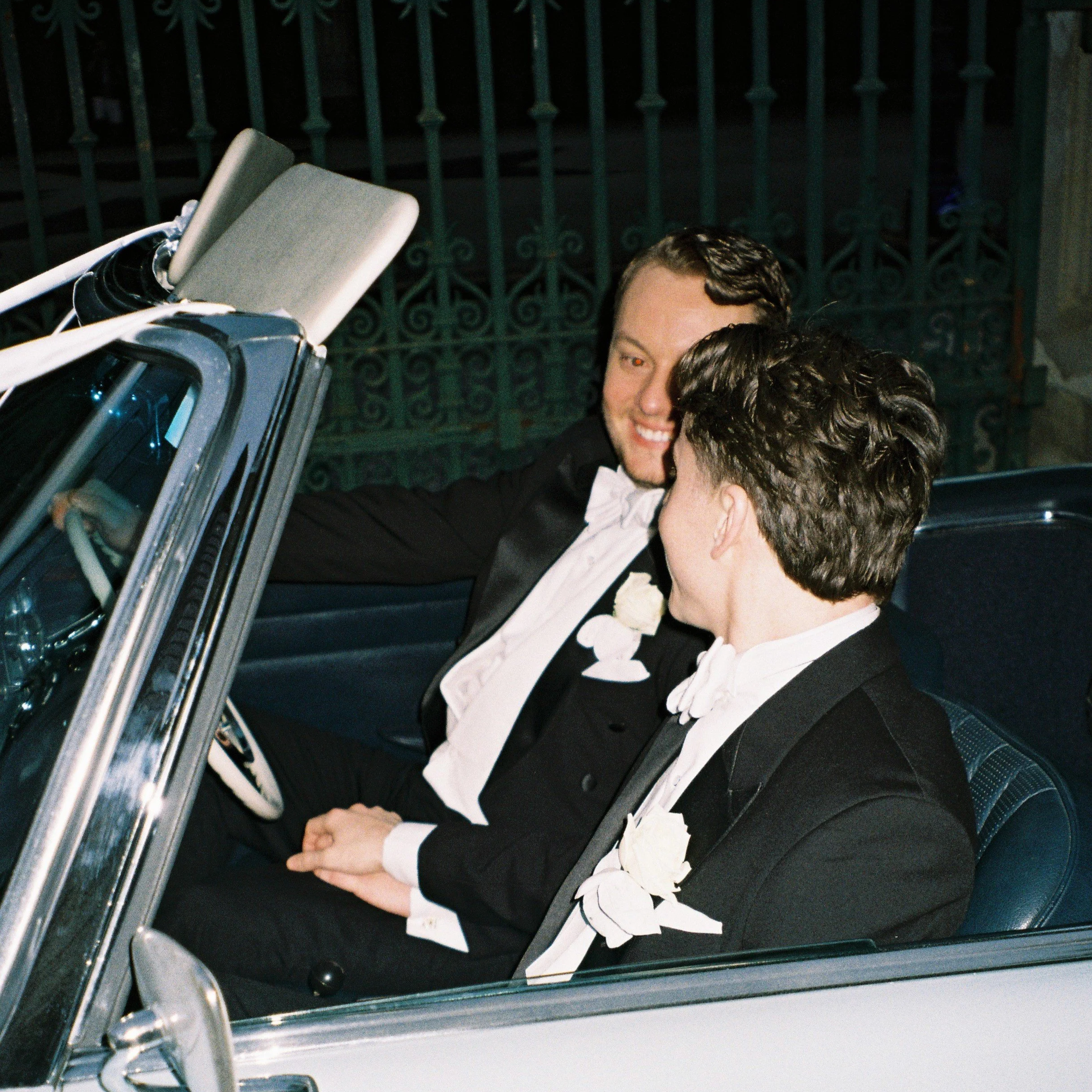 Two men in tuxedos embrace each other in a close hug outdoors at night near a green wrought iron fence, with a vehicle nearby.