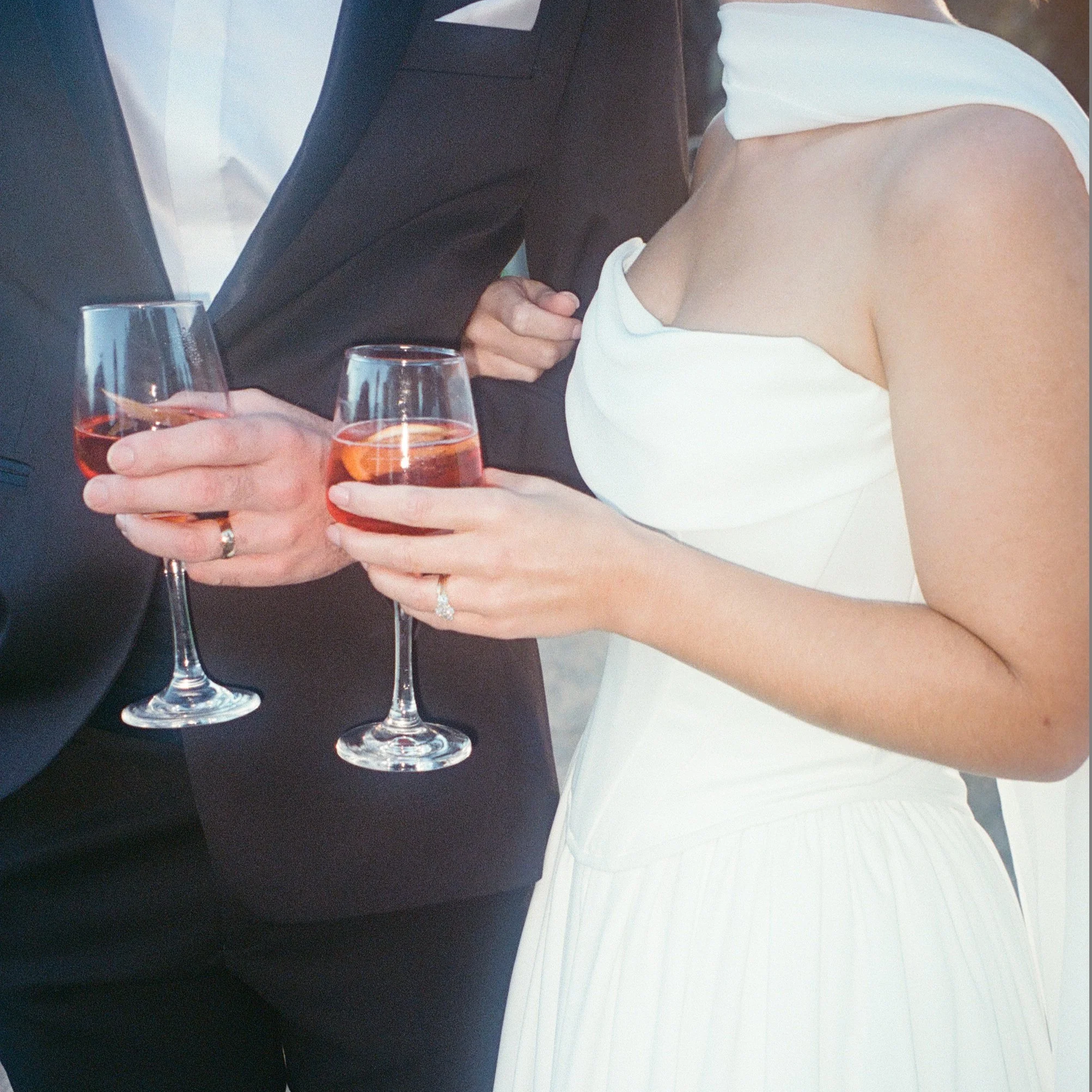 A couple holding glasses of rosé wine, with the woman's right hand wearing a diamond ring, in a setting with a black table and possibly a white or light-colored dress.