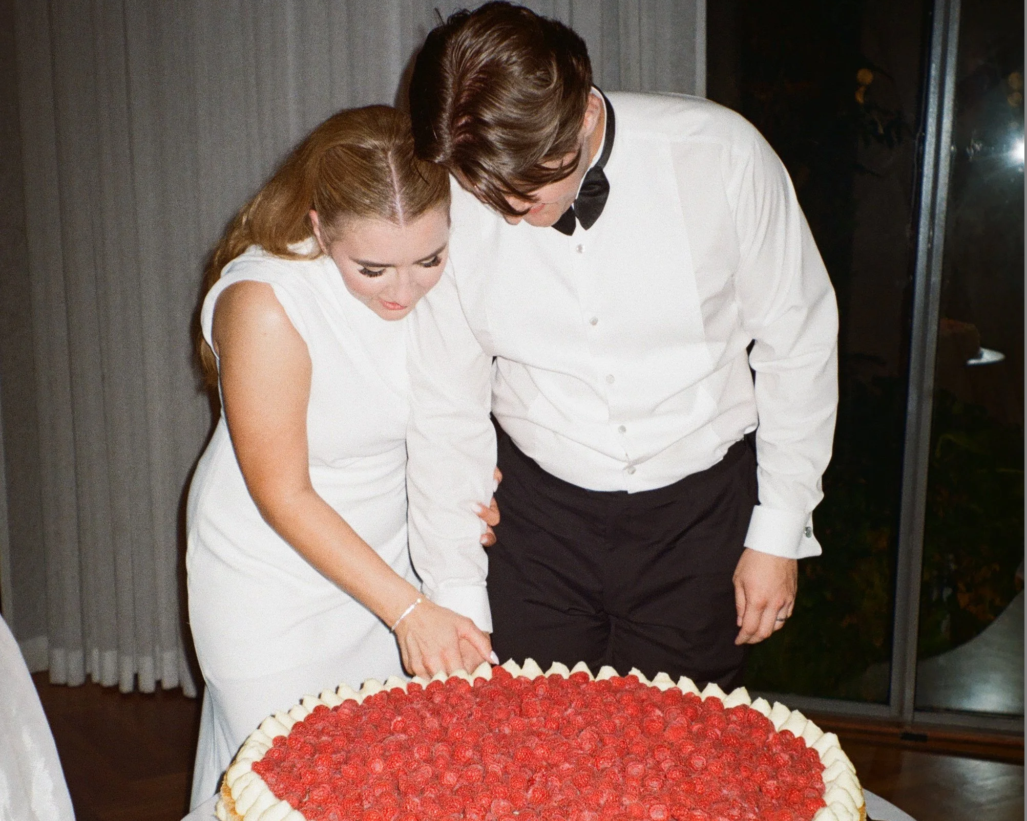 A couple, dressed in formal attire, is cutting a large raspberry-topped cake together at an event.