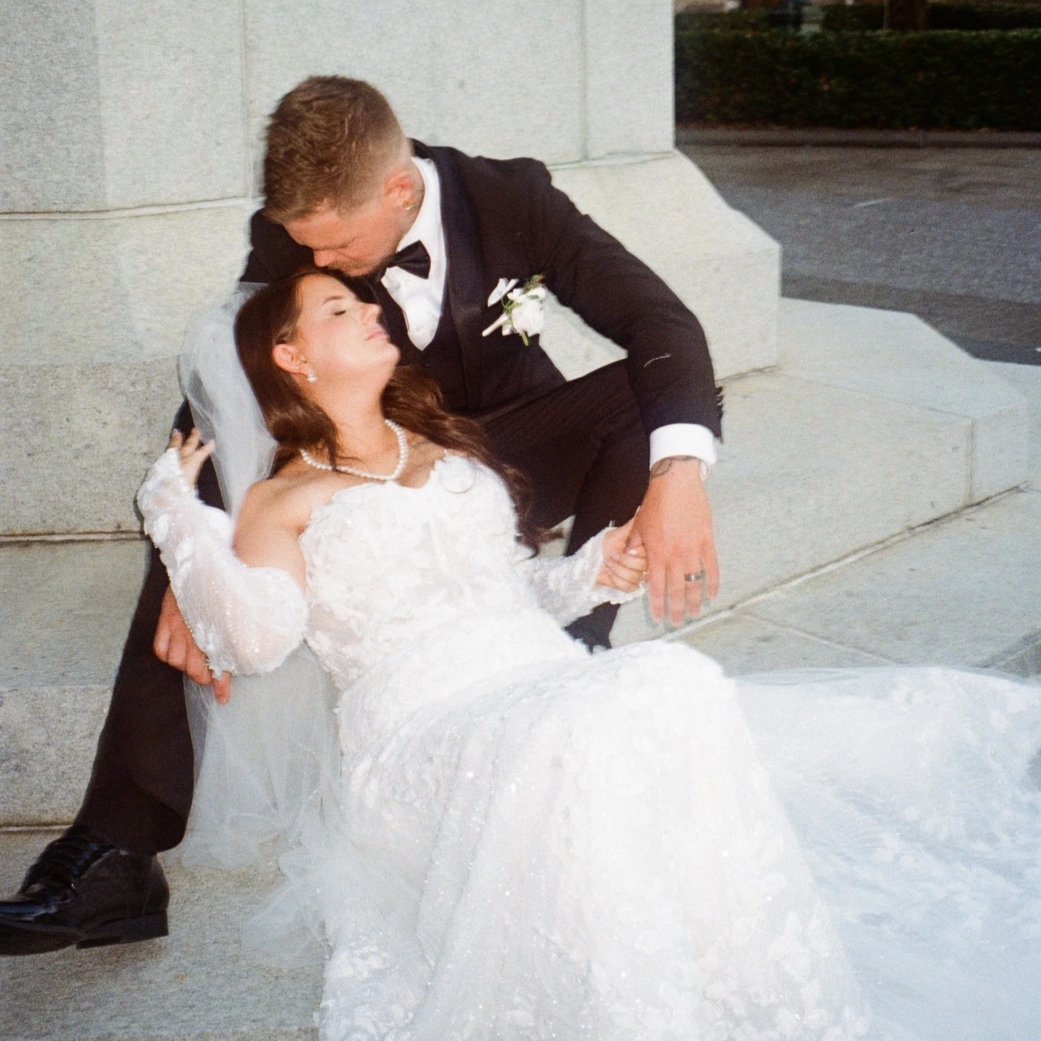 A groom in a tuxedo kissing a bride in a white wedding dress and pearl necklace, lying on the ground against a gray wall, with an outdoor setting visible at night.