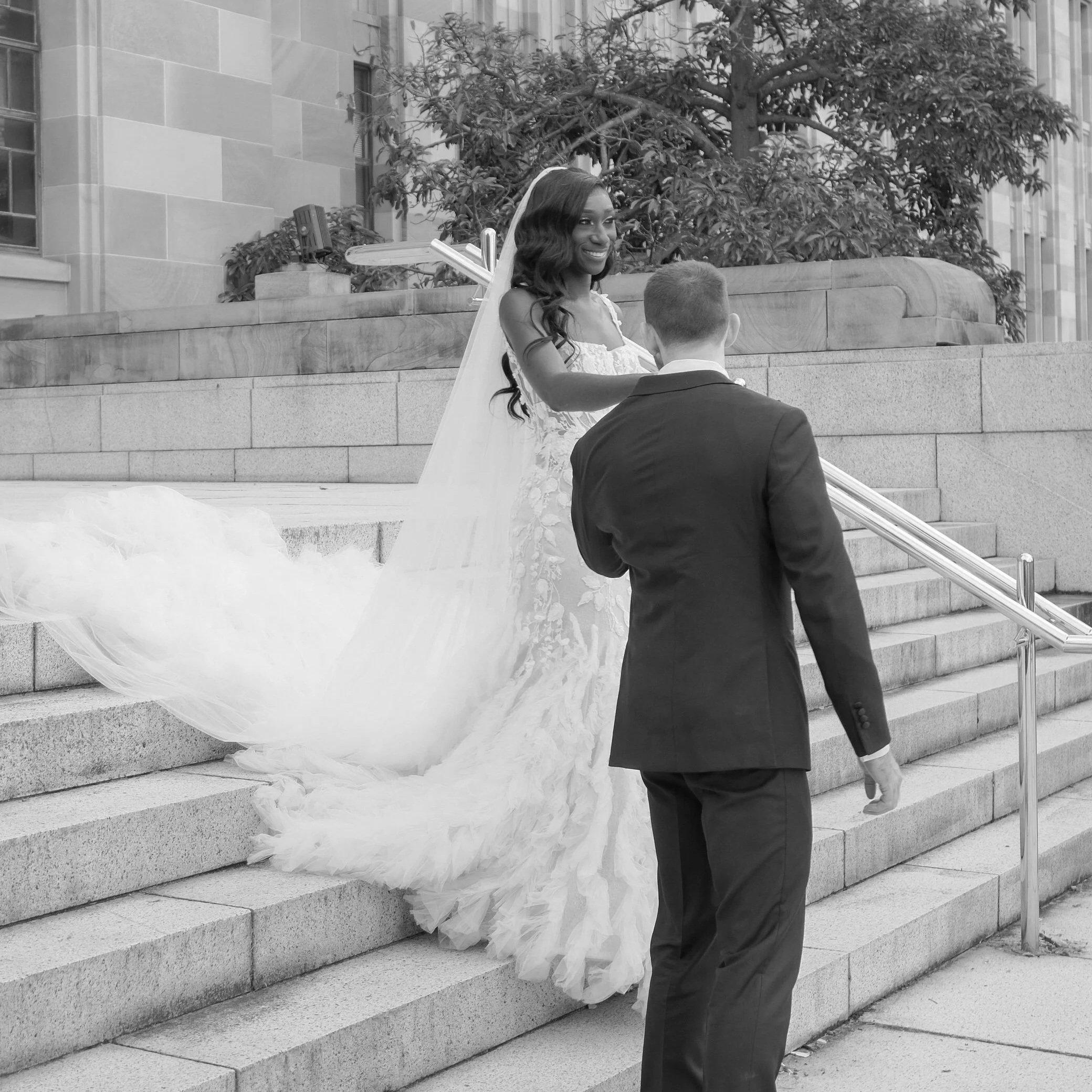 A woman in a wedding dress and veil smiling at a man in a suit, standing on outdoor stairs with a large tree and a building in the background, during a wedding ceremony.