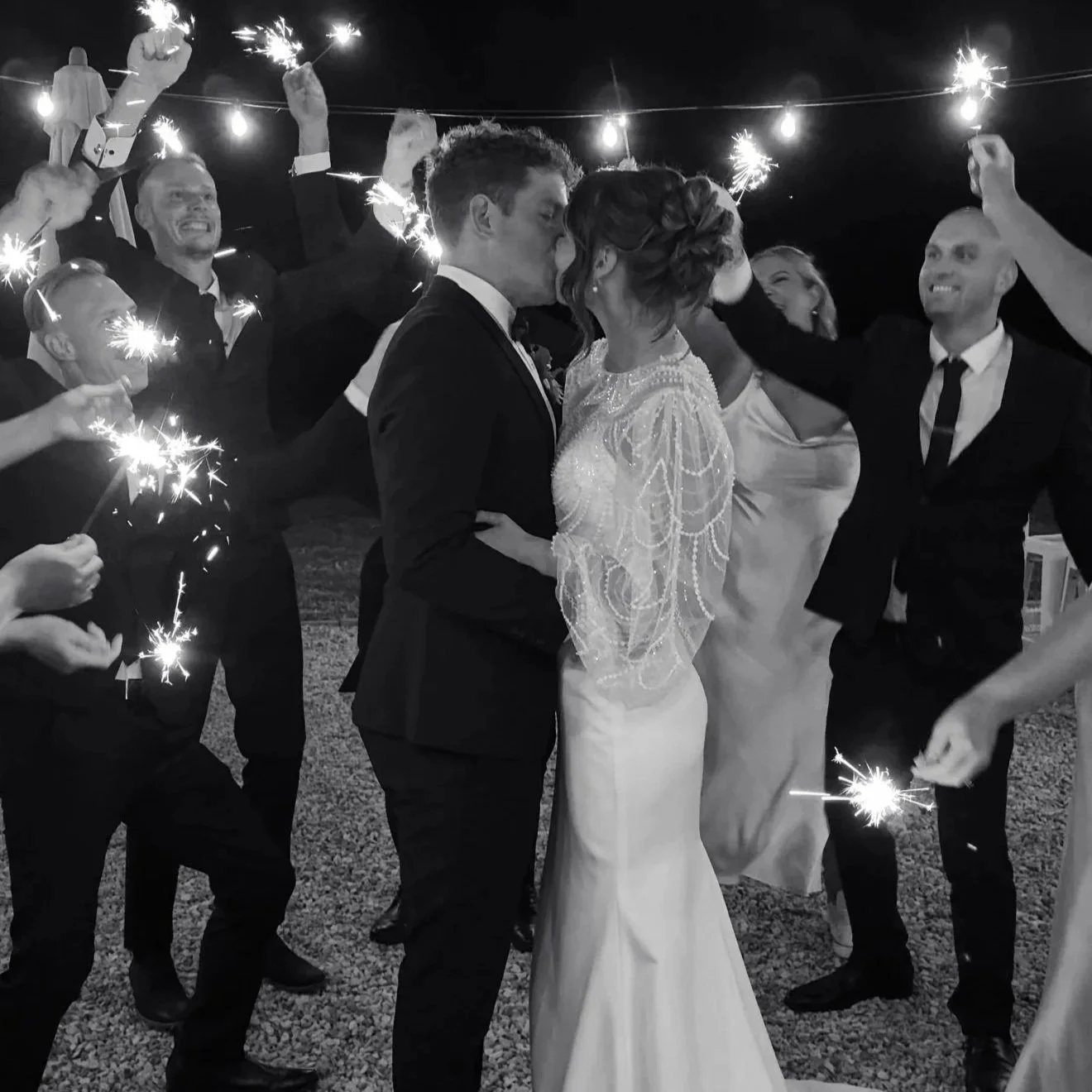 Black and white photo of a wedding celebration with a bride and groom kissing surrounded by friends holding sparklers.