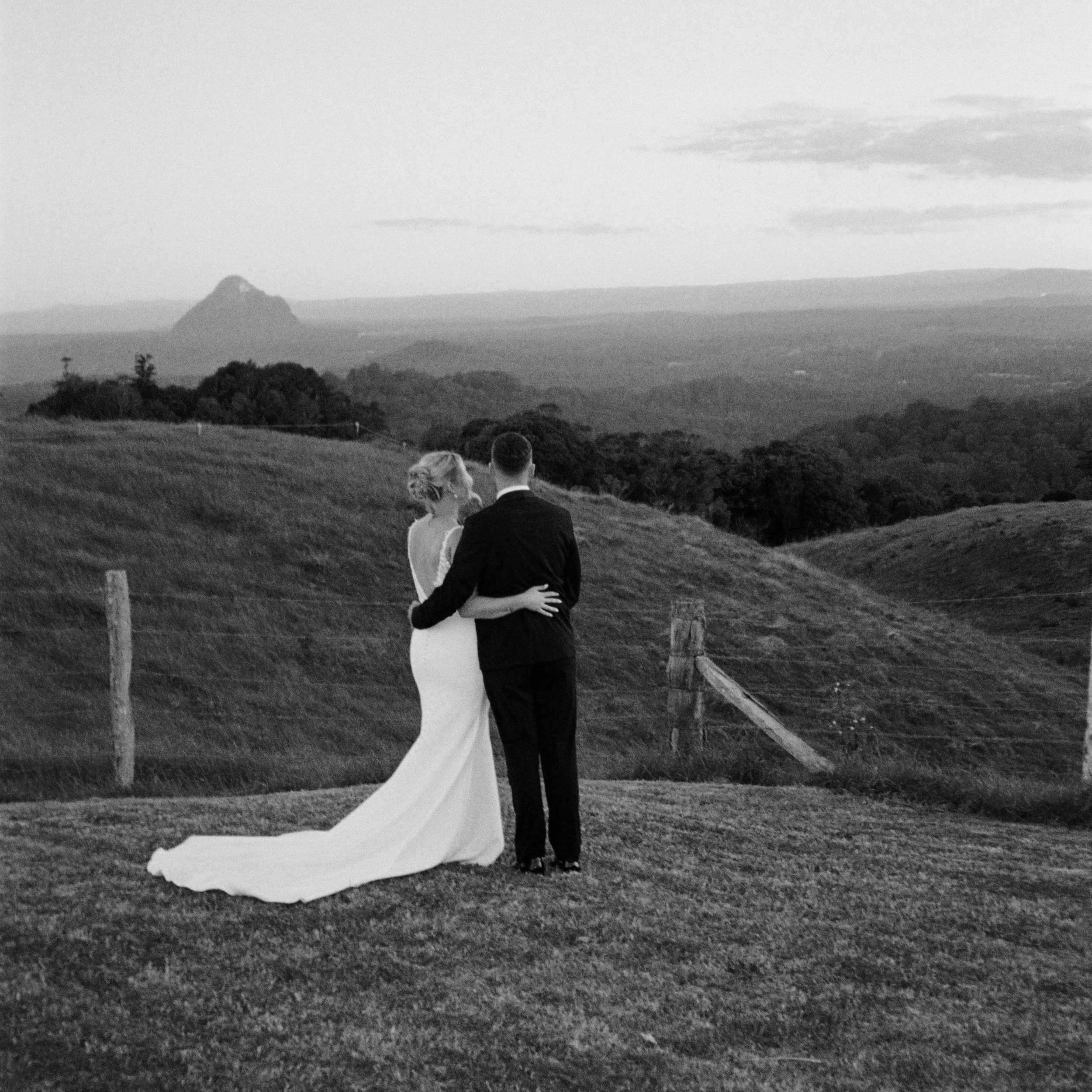A couple, dressed in wedding attire, standing on a grassy hill, holding each other and looking at a scenic landscape with rolling hills and a mountain in the background.