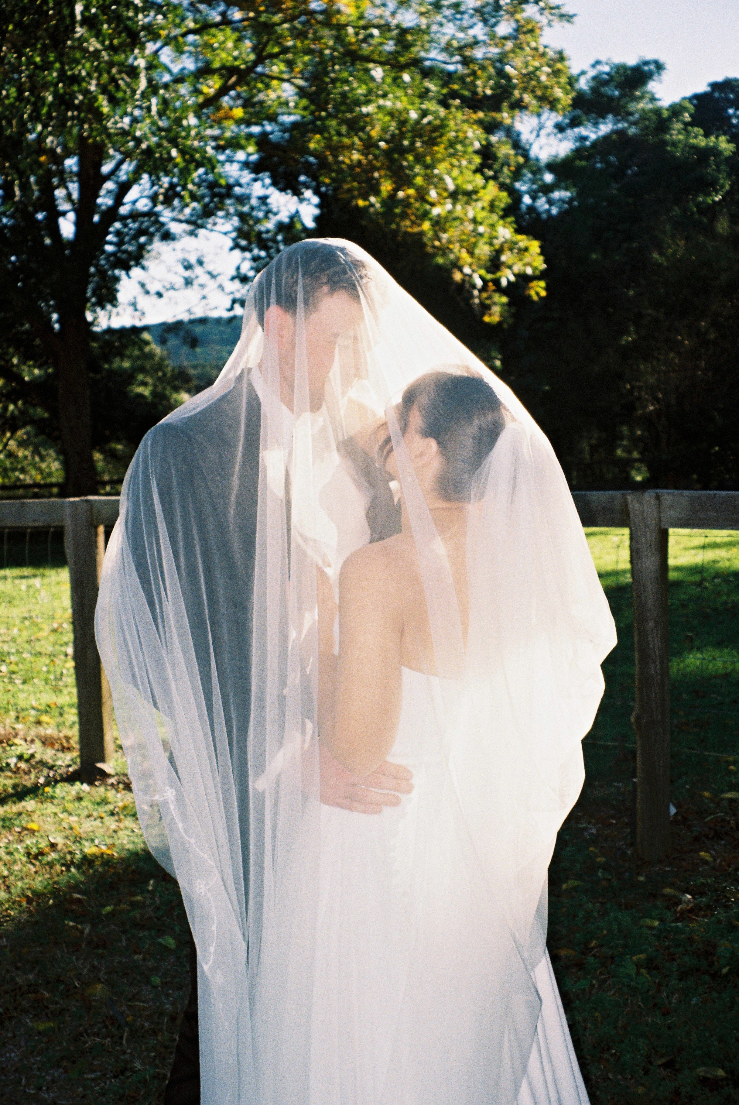 A bride and groom outdoors under a veil, sharing a tender moment with trees and a wooden fence in the background.