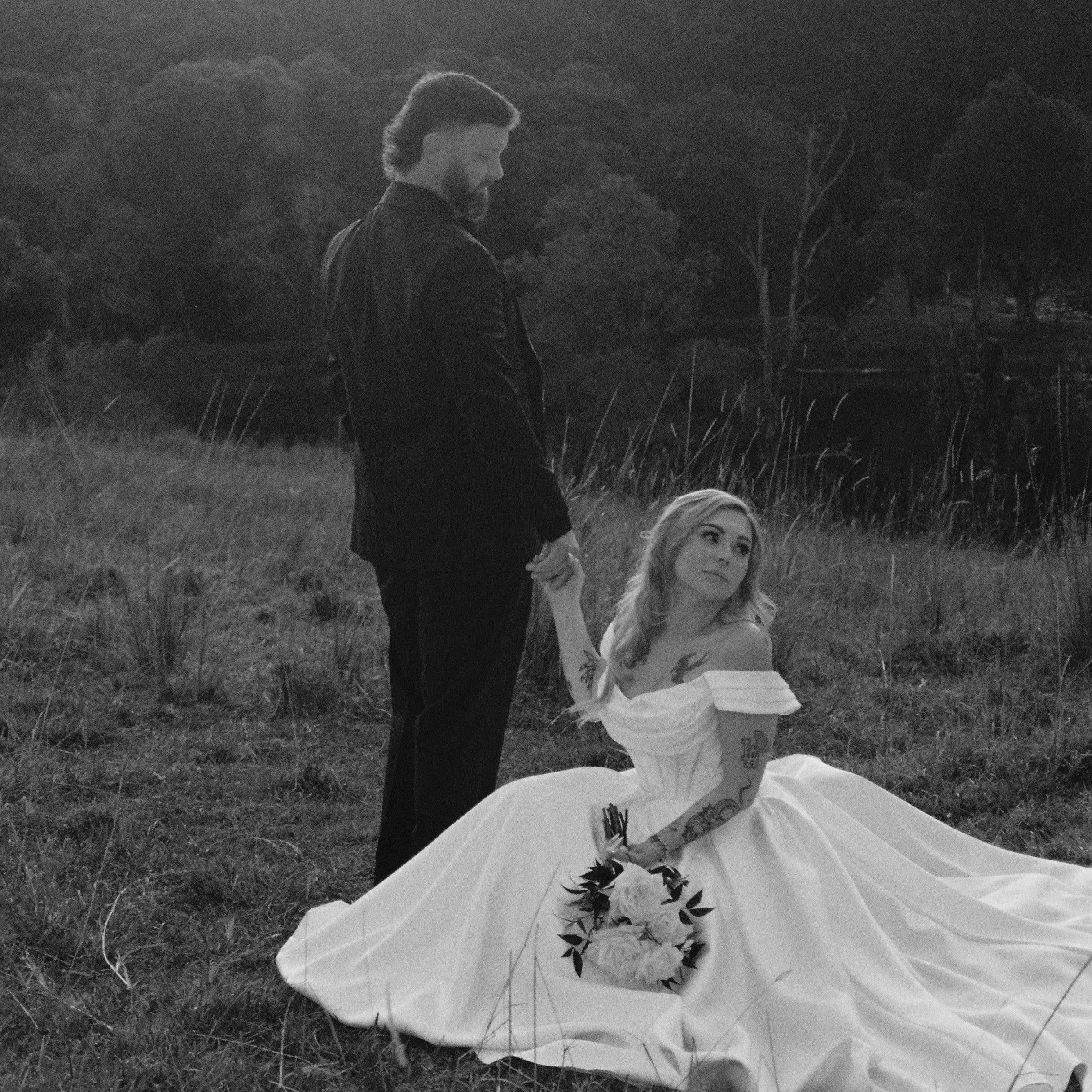 A man with a beard and long hair standing outdoors at dusk or dawn, holding a woman's hand, with trees and grass in the background.