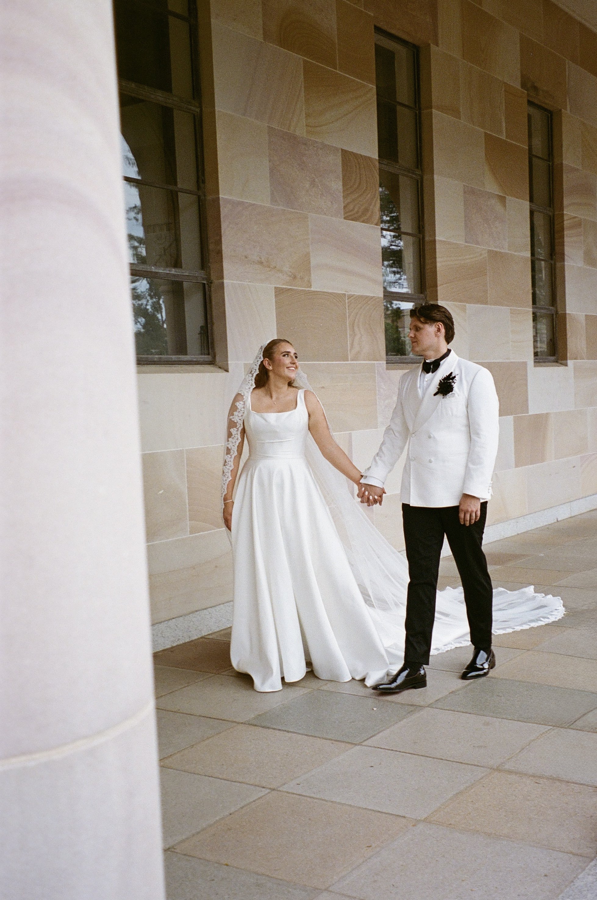 A bride and groom holding hands outside a building with large windows and stone walls.
