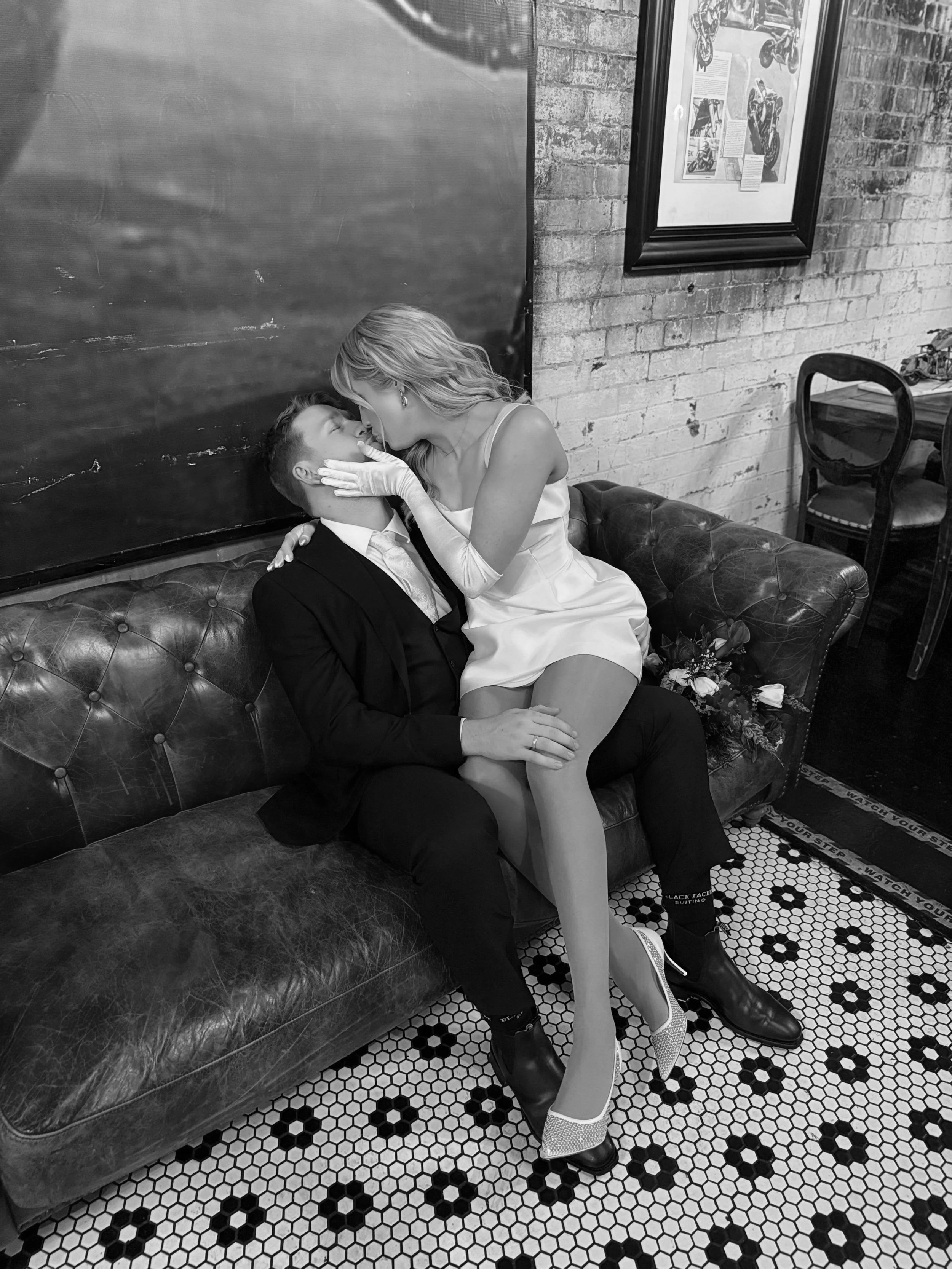 A black and white photo of a woman in a dress and heels leaning over a man in a suit sitting on a leather couch, sharing a kiss in a room with brick walls, framed pictures, and tiled flooring.