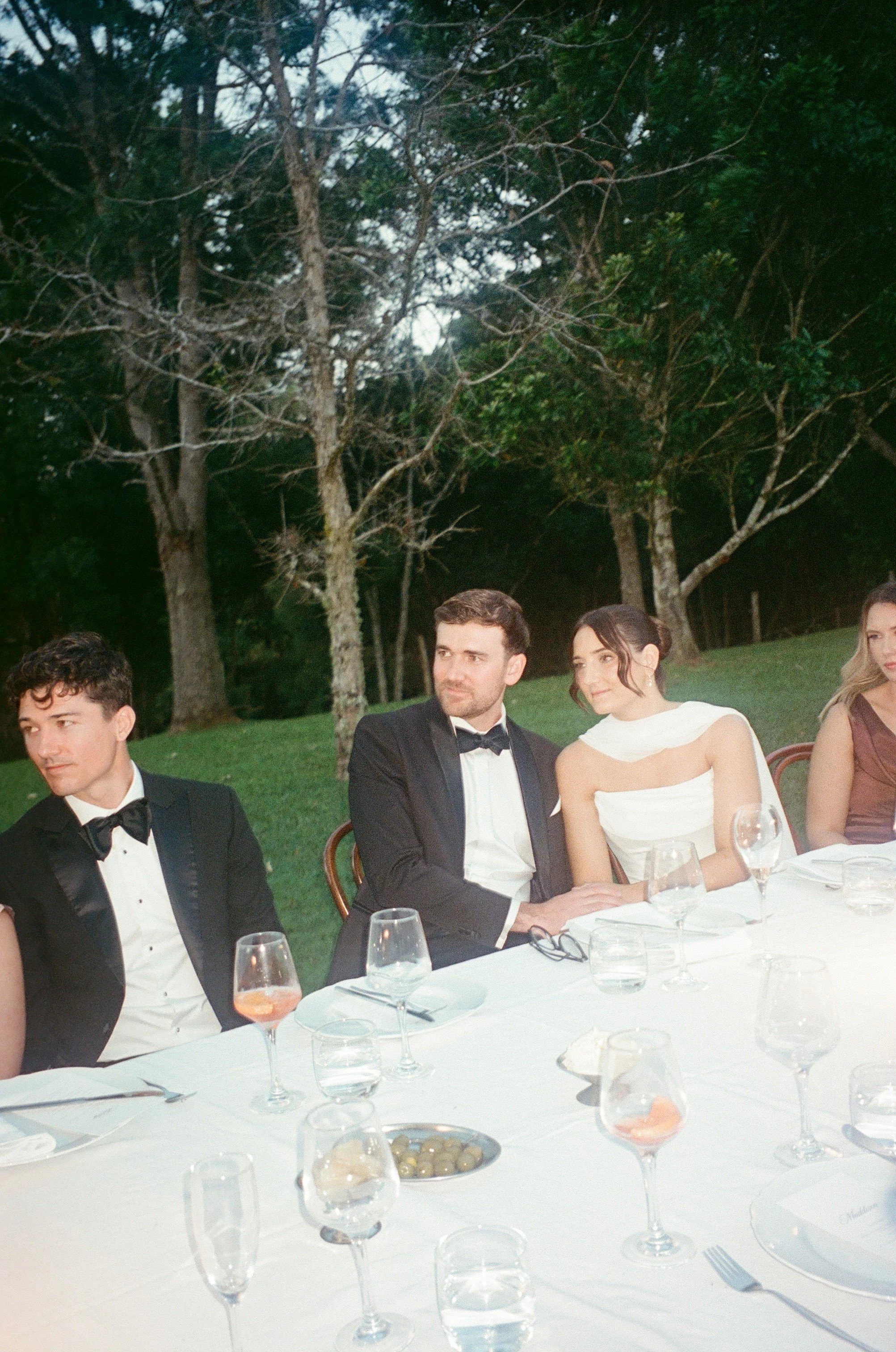 People dressed in formal attire sitting at a round table outdoors during an event or celebration, with a wooded background.
