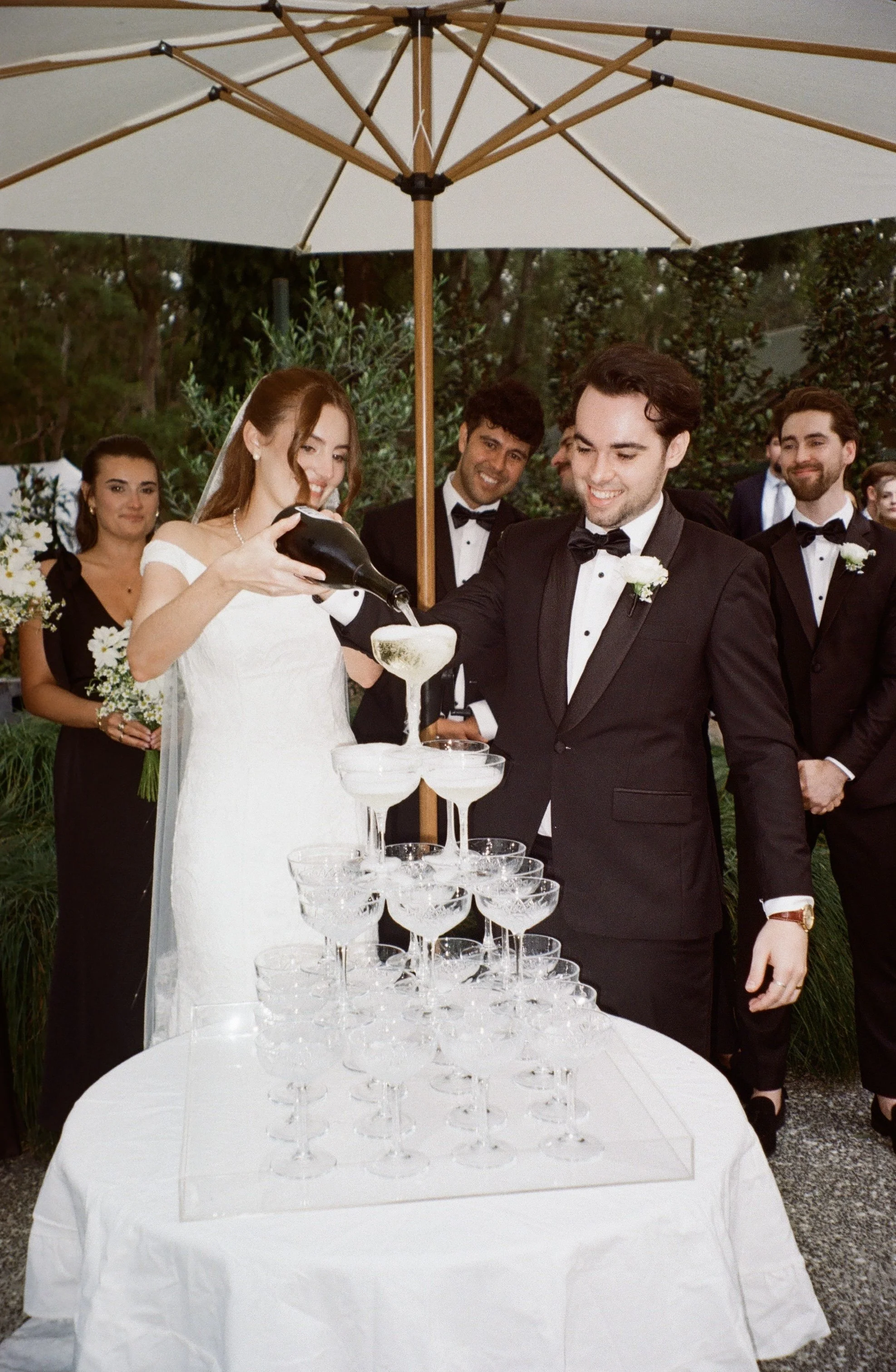 A wedding celebration outdoors with the bride and groom pouring champagne into a tower of glasses, surrounded by bridesmaids and groomsmen in formal attire, under a large white umbrella with trees in the background.