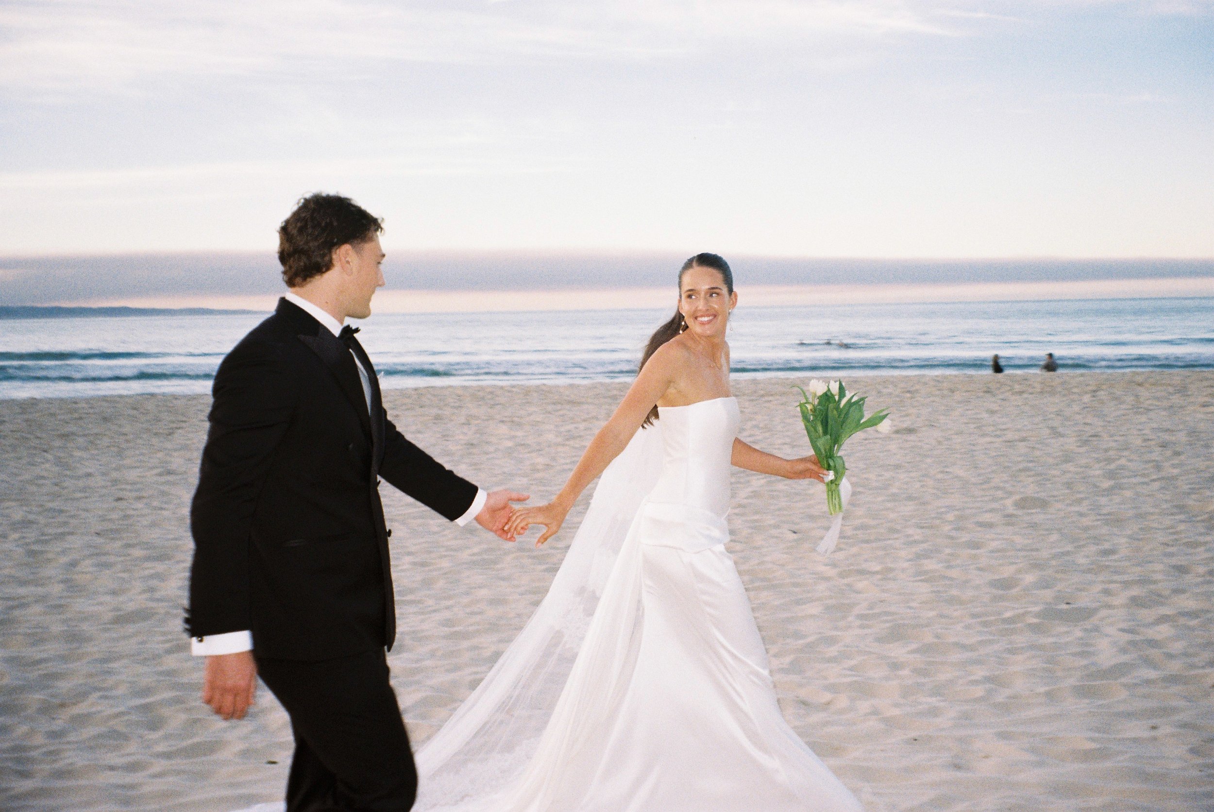 A bride and groom hold hands and walk on the beach during their wedding, with the ocean and sky in the background, and the bride carrying a bouquet of flowers.