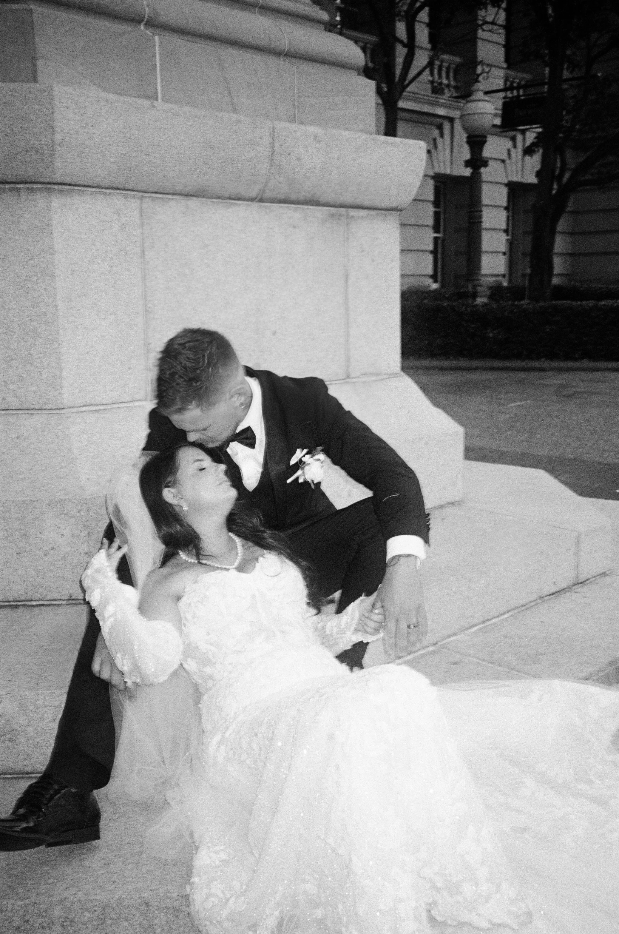 A bride and groom dance together outdoors at night, with the groom dipping the bride in front of a stone building and street lamps.