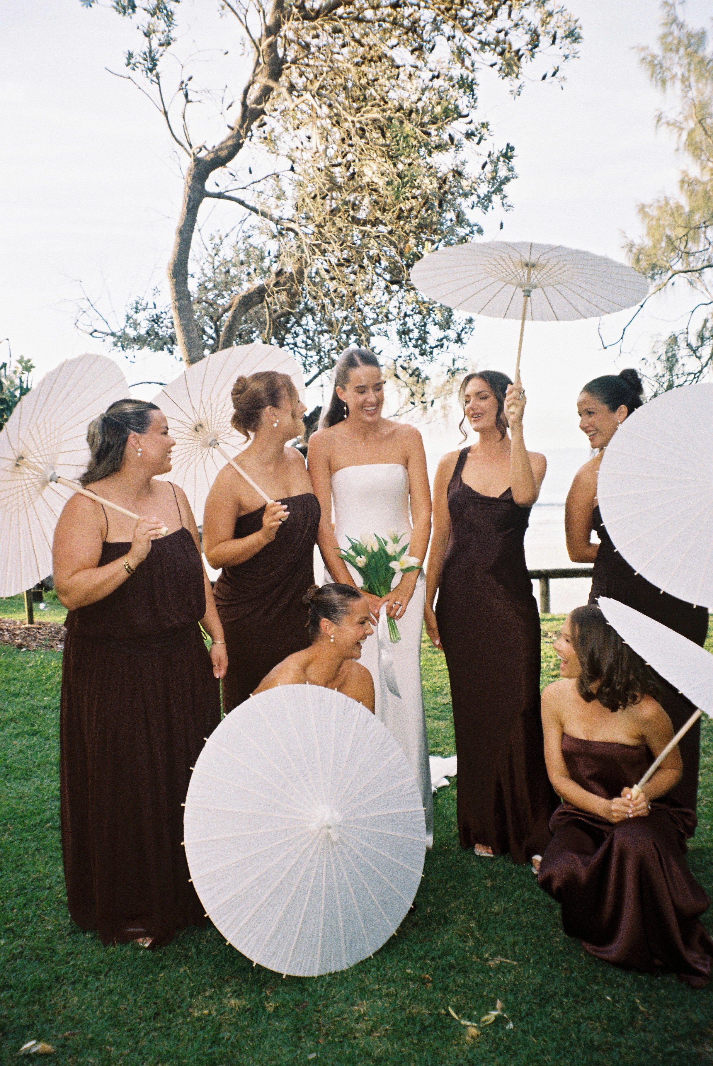 A group of women dressed in formal dresses, some holding white parasols, standing outdoors on a grassy area with trees in the background.