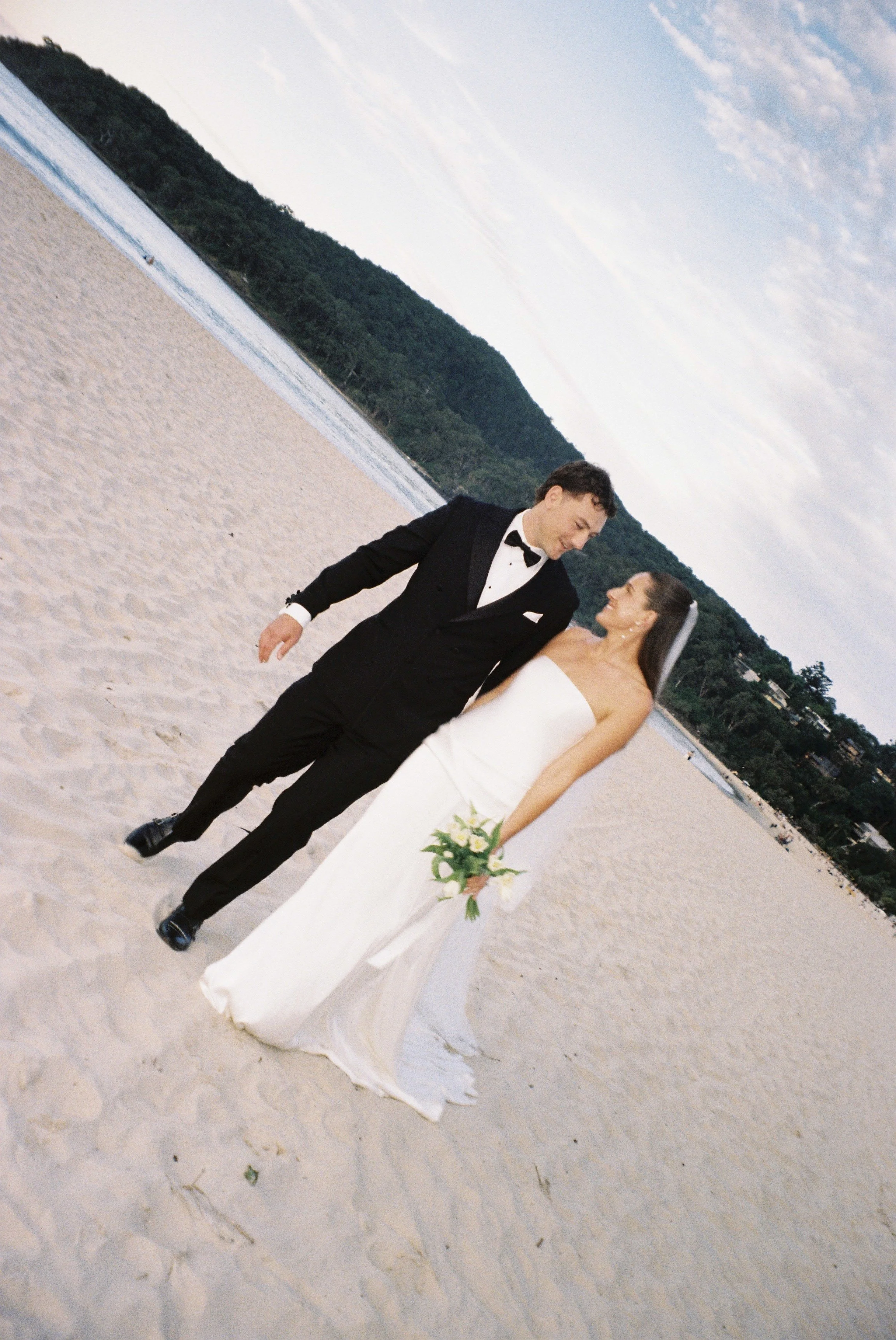 A newlywed couple walking on a sandy beach with a body of water and a forested hill in the background during sunset or early evening.