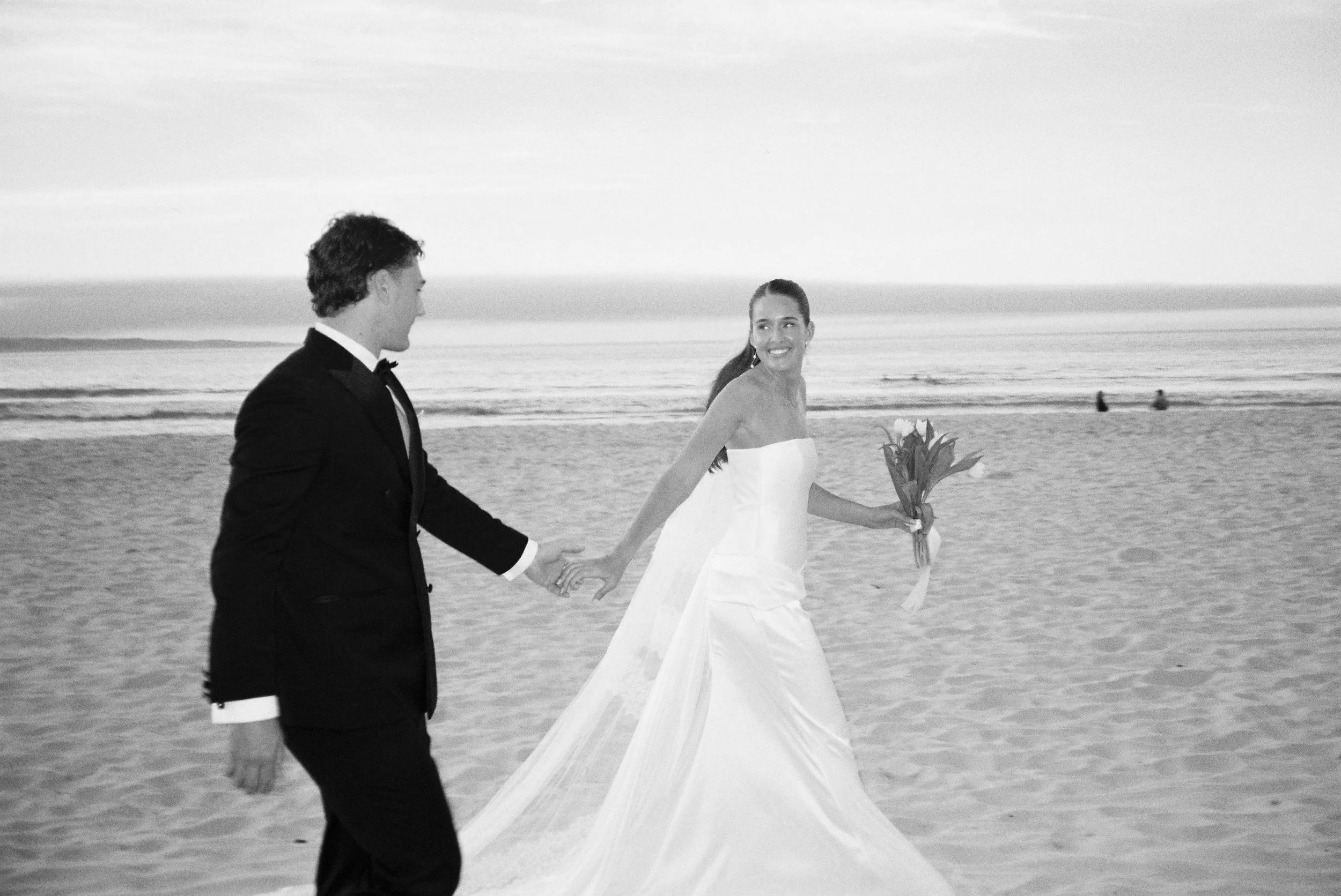 A couple holding hands and walking on the beach, with the woman in a wedding dress holding a bouquet of flowers, and the man in a tuxedo, during sunset or sunrise.