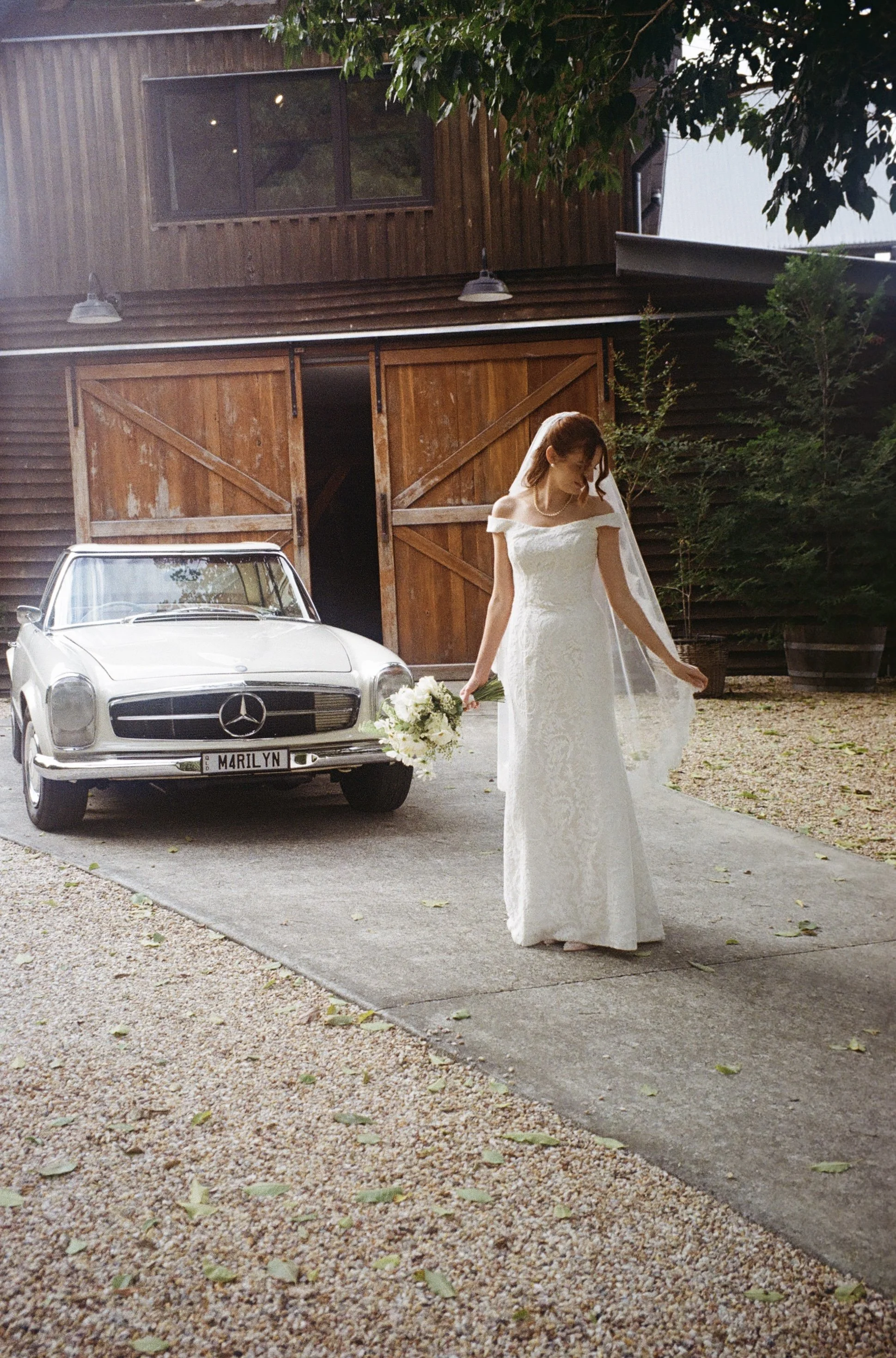 A woman in a wedding dress holding a bouquet in front of a wooden barn with a vintage Mercedes car parked nearby.