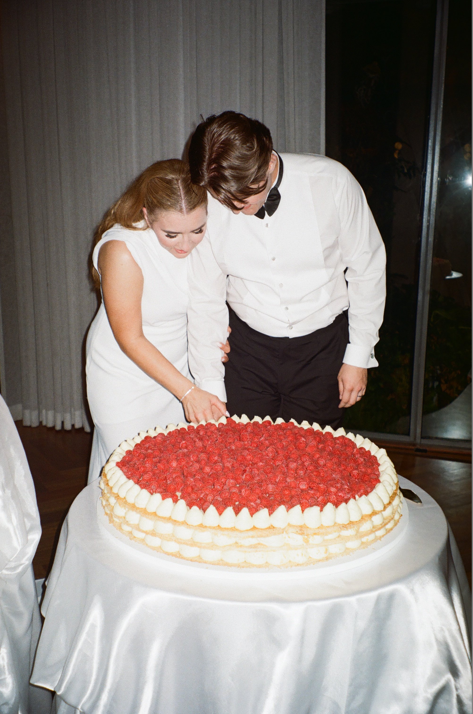 A man and woman dressed in formal attire cutting a large wedding cake decorated with raspberries and piped cream.