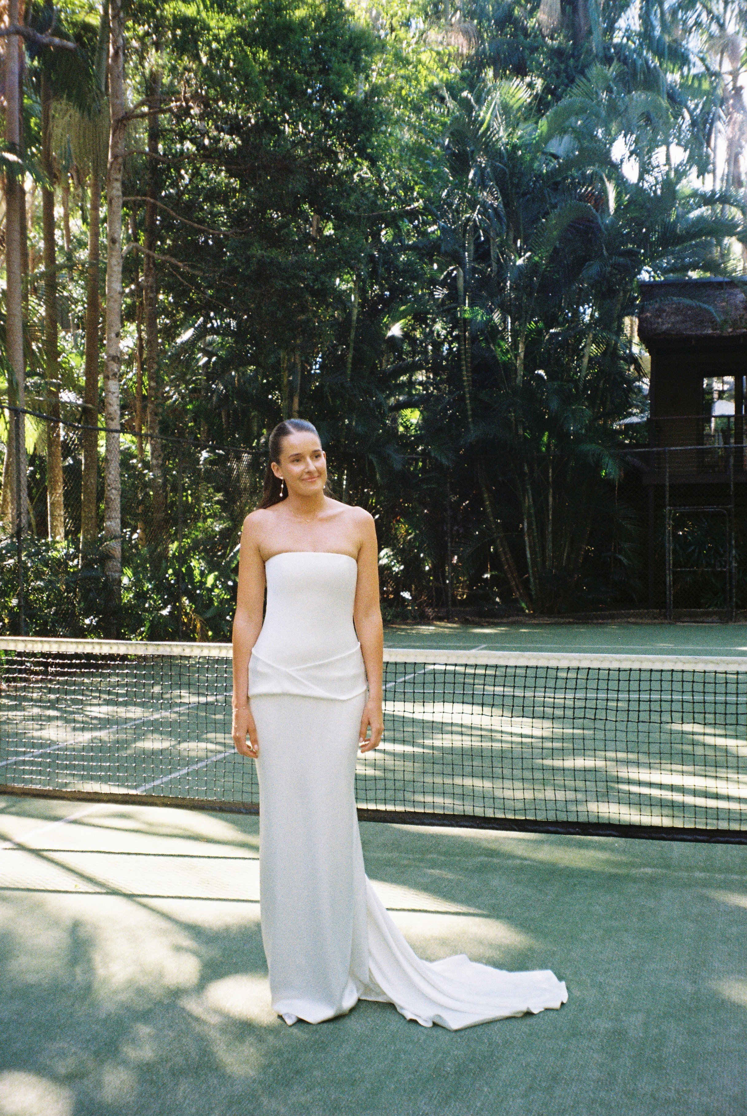 A woman in a strapless white gown standing on a tennis court with a net in front of her, surrounded by lush green trees.