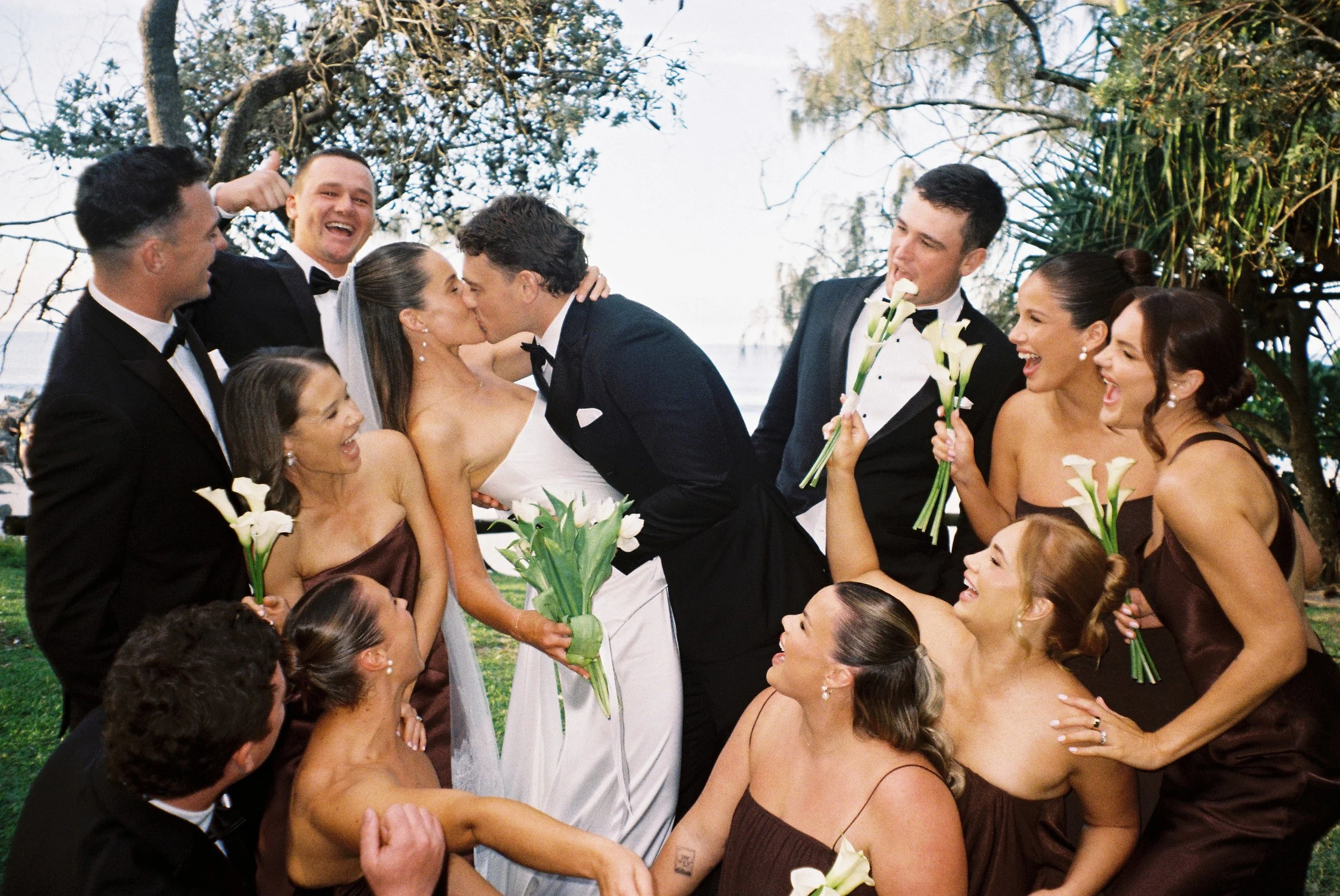 A group of men and women at a wedding, with two men and a woman kissing in a circle of smiling friends holding calla lilies. The setting is outdoors with trees and sky in the background.