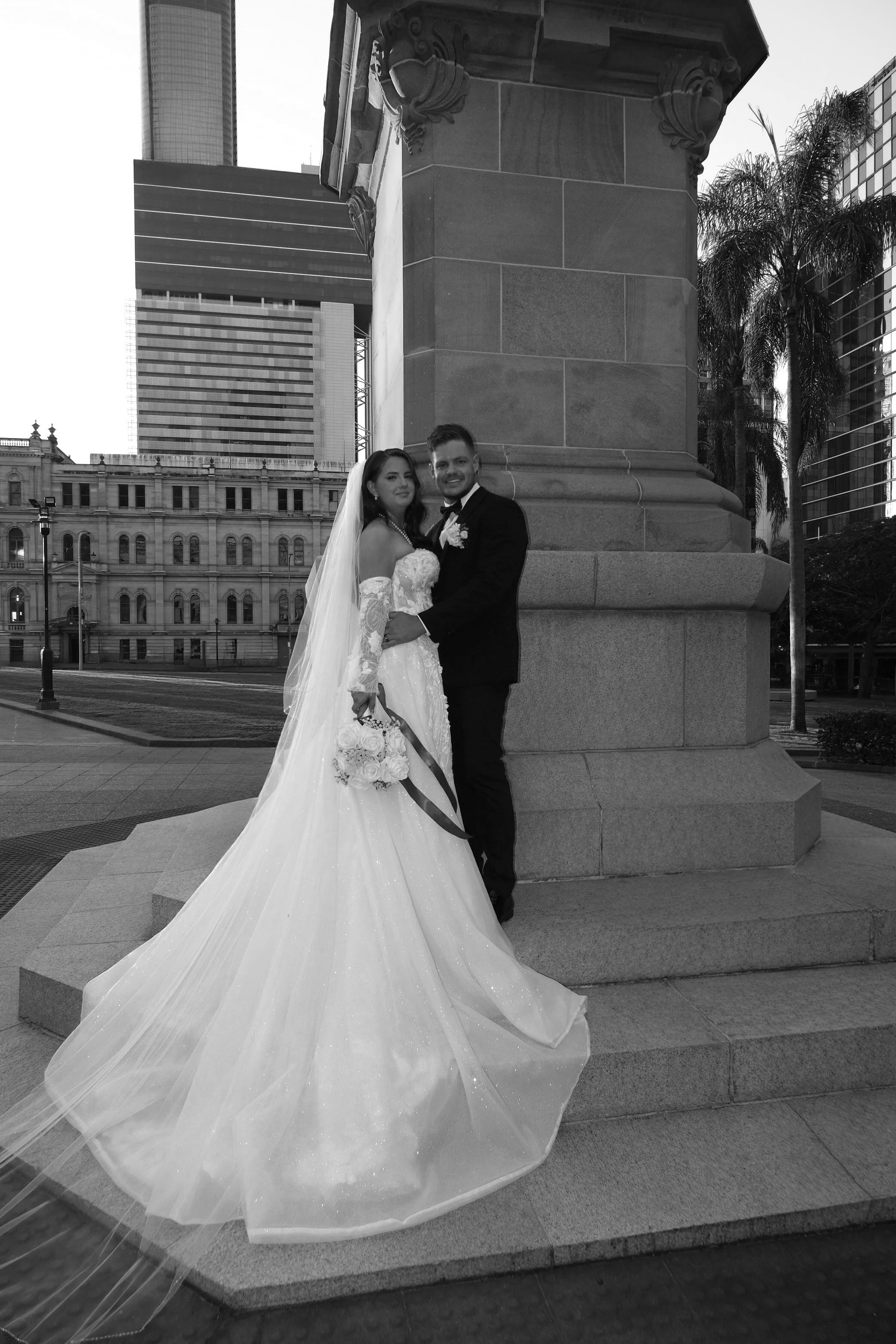 A black and white photo of a newlywed couple standing outdoors at the base of a large stone monument. The bride is wearing a wedding gown with lace detail and holding a bouquet of flowers. The groom is in a tuxedo, smiling while standing close to the bride. Tall buildings and palm trees are visible in the background.