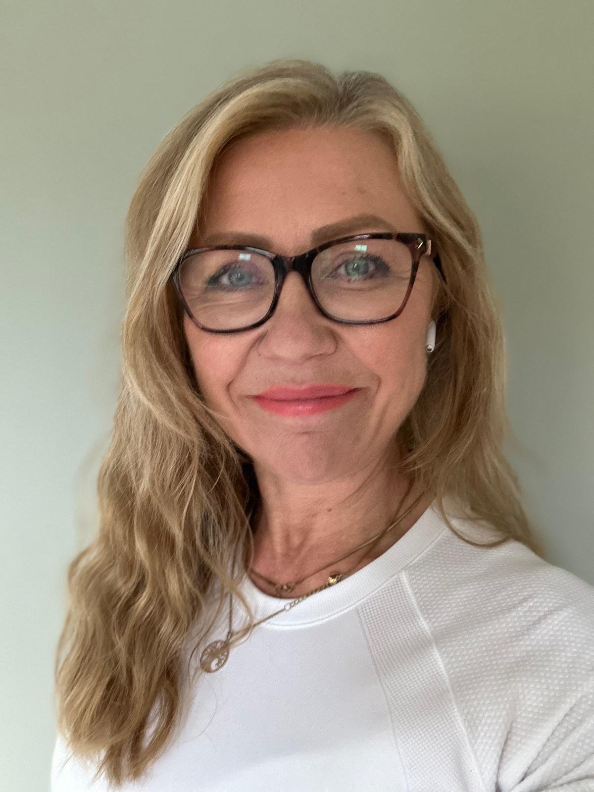Jane Wrafter Counselling,  with long wavy blonde hair, wearing glasses, a white top, and a necklace, smiling against a plain green wall.