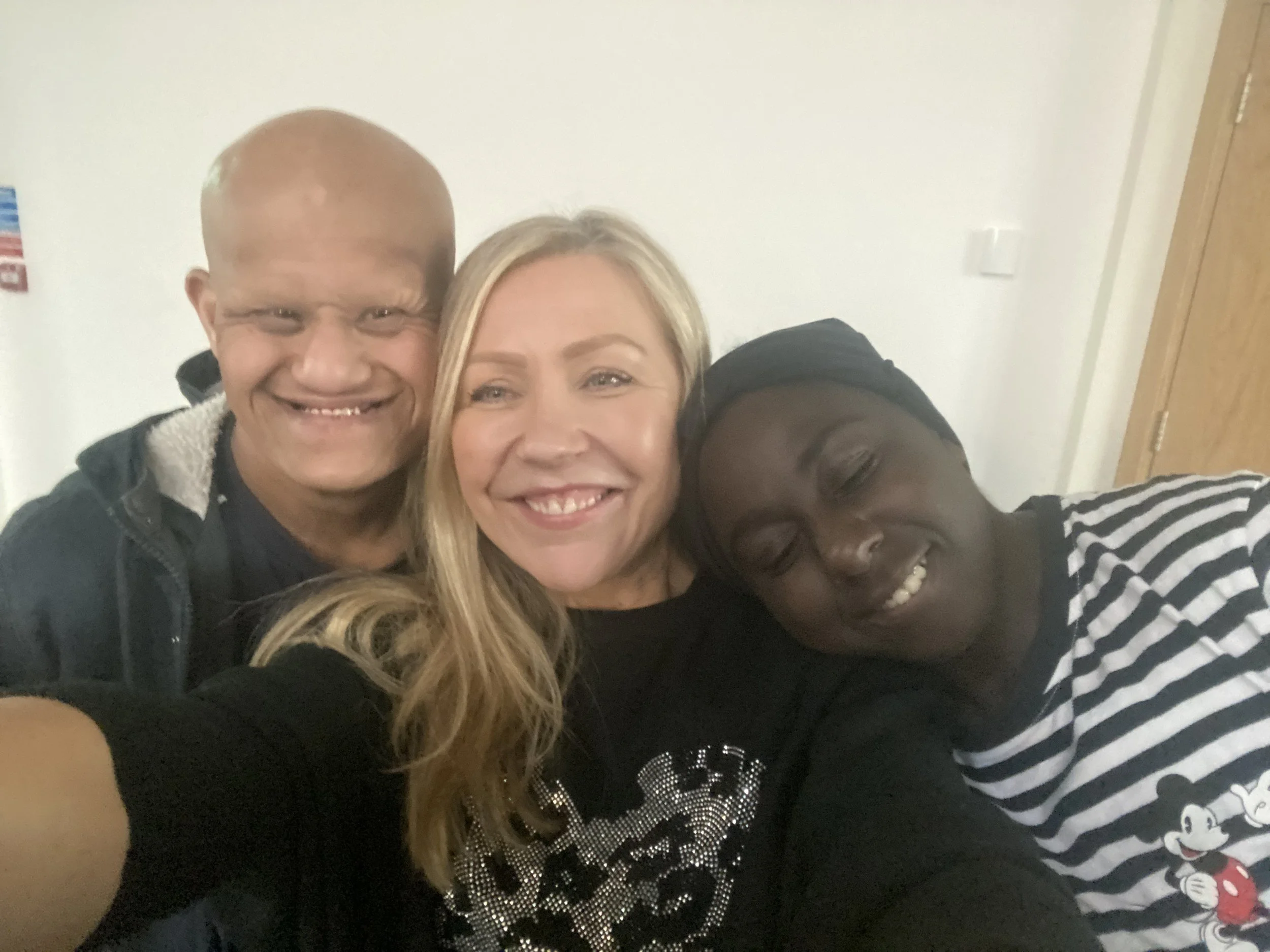 Three people smiling and leaning close together for a selfie, indoors with a plain wall in the background.